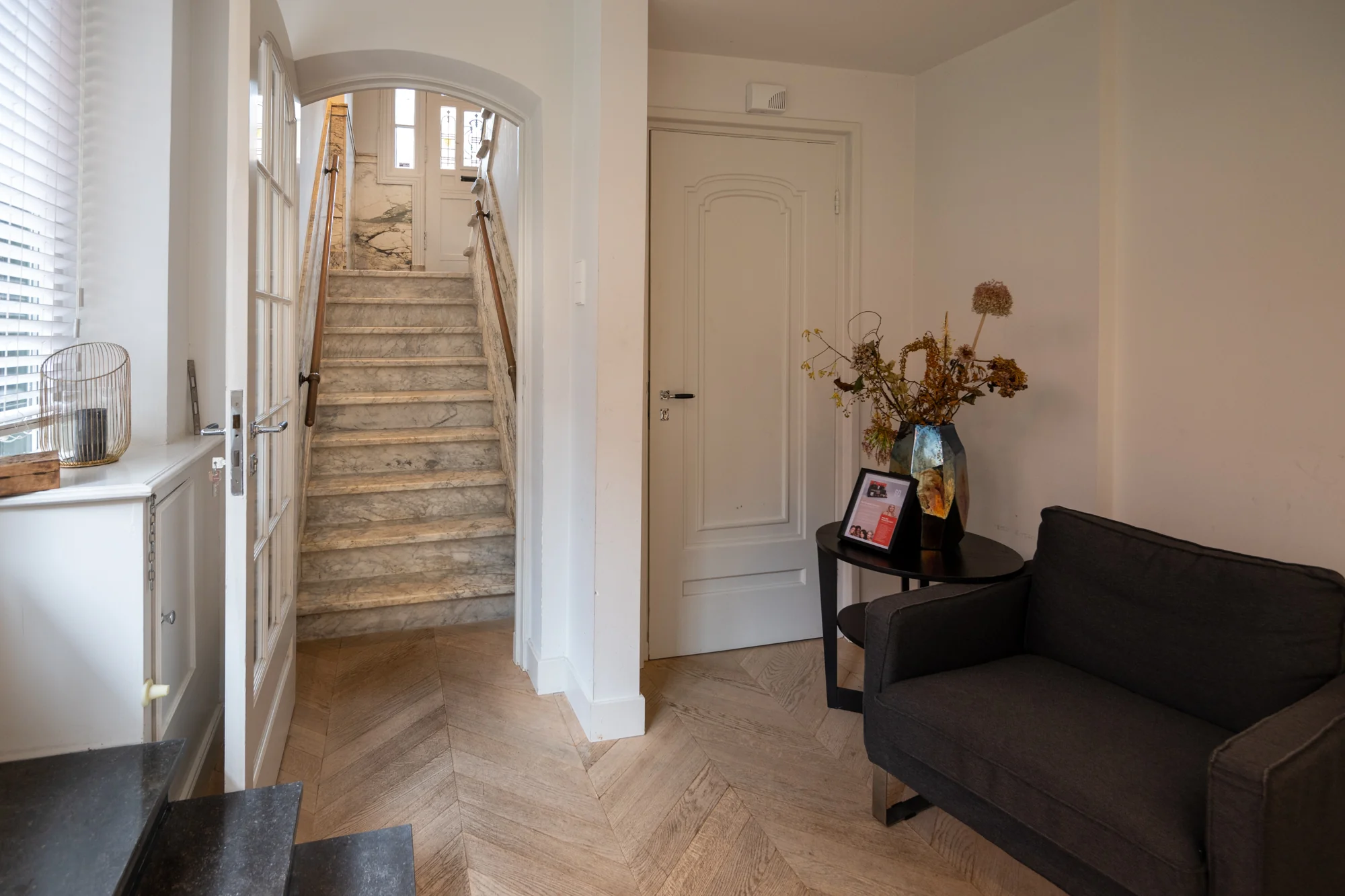 Hal met marmeren trap, houten visgraatvloer, zwarte fauteuil en een bijzettafel met vaas en ingelijste foto in een woning aan de Hobbemastraat.