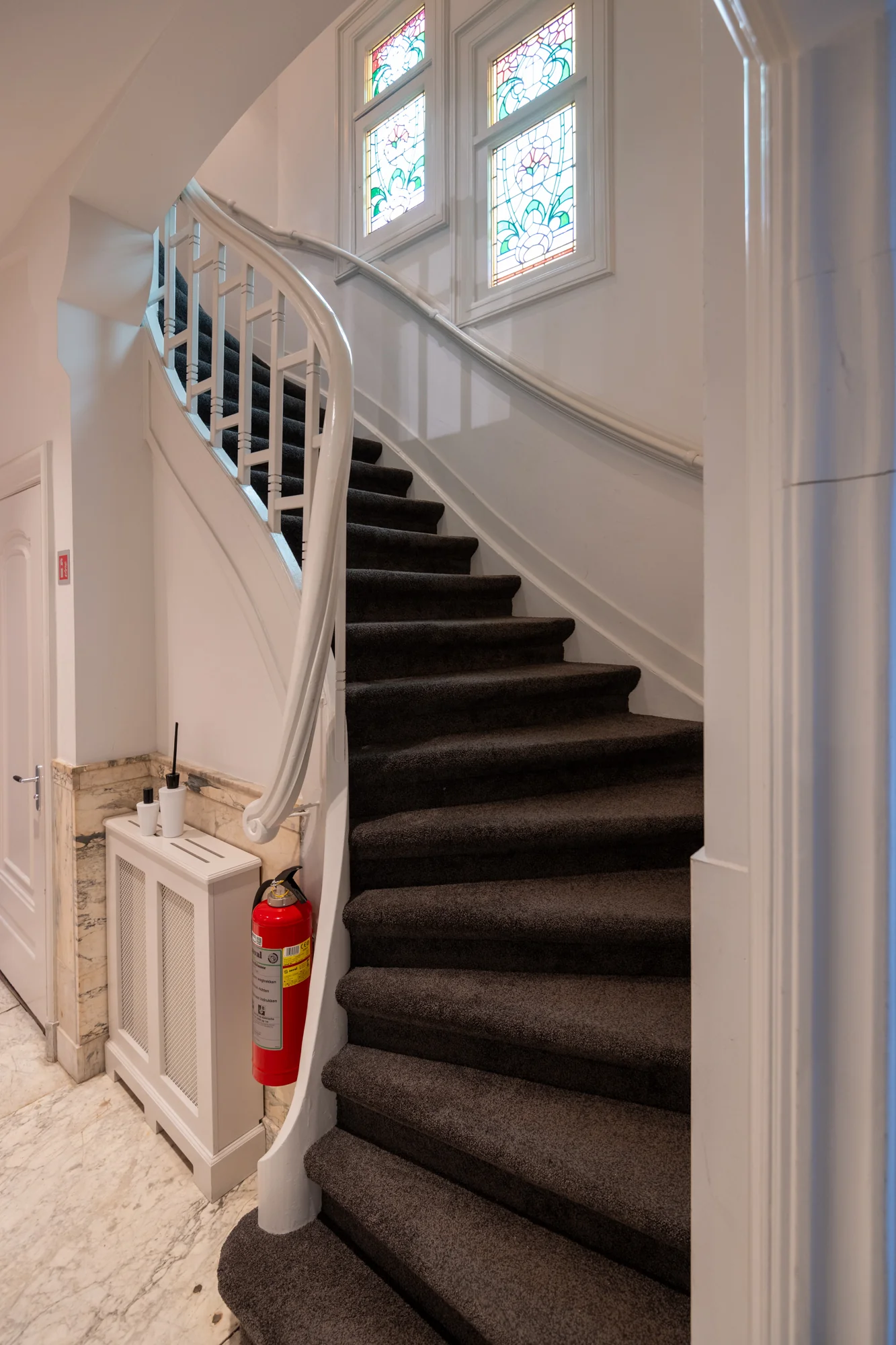 Wenteltrap met donker tapijt en wit houten leuning, naast glas-in-loodramen in een pand aan de Hobbemastraat.