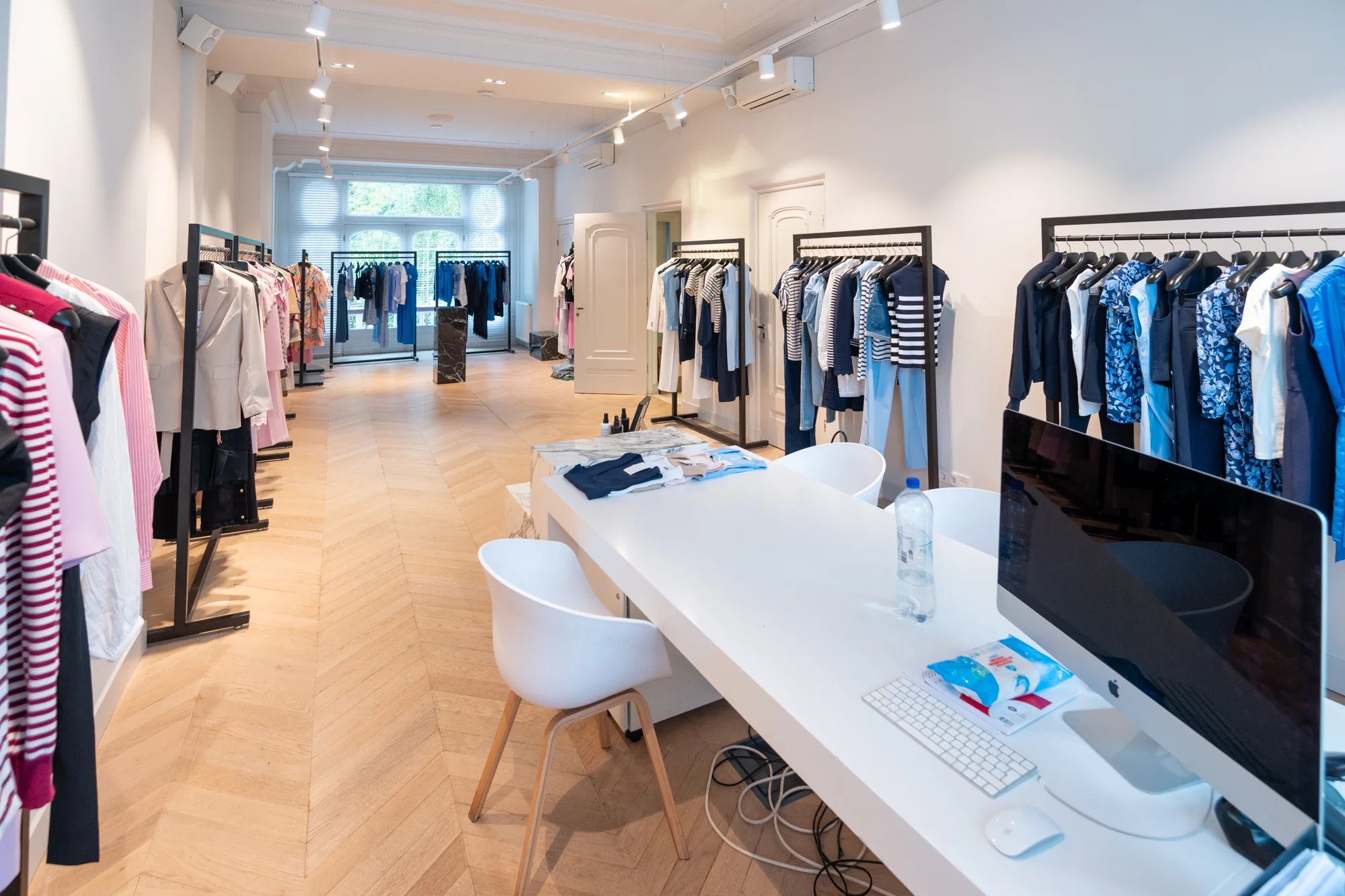 Moderne showroom met kledingrekken langs de wanden en een witte werktafel met computer in het midden op de Hobbemastraat.