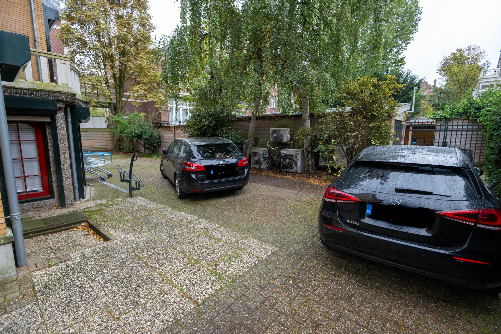 Twee geparkeerde auto's op een natte binnenplaats aan de Hobbemastraat, omgeven door bomen en gebouwen.