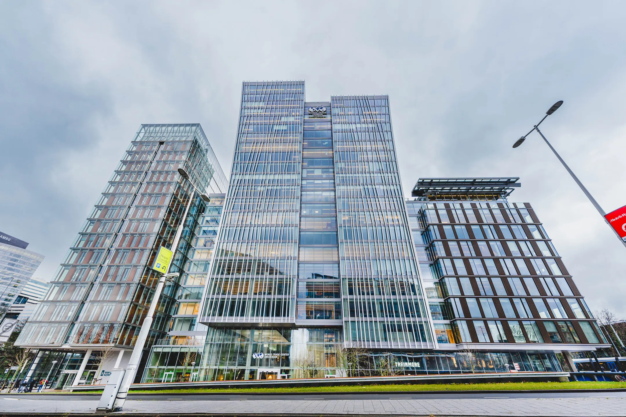 Een moderne, glazen kantoorbouw in een stedelijke omgeving onder een bewolkte hemel.