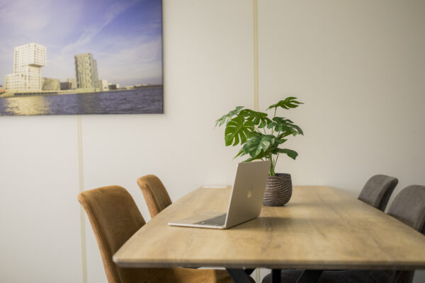 Een houten vergadertafel met een laptop en een plant erop in een modern kantoor, met een foto van een architectonisch gebouw aan de muur.