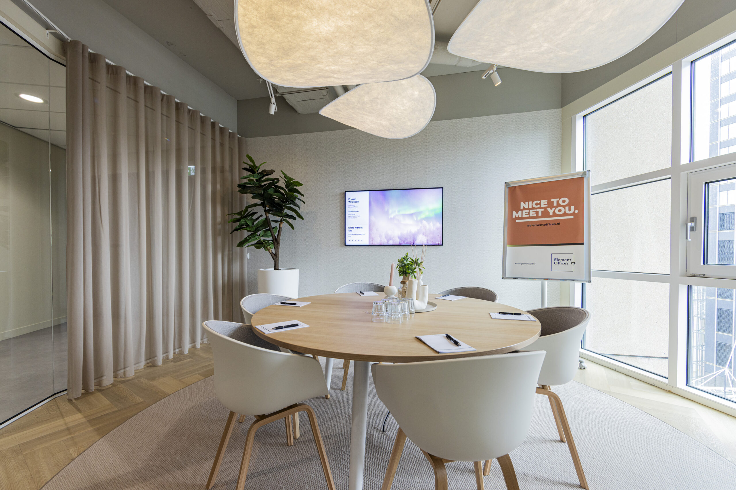 Een moderne vergaderruimte met een ronde tafel, stoelen, een plant, een scherm en een welkomstbord.