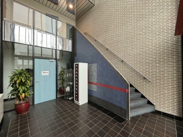 Ingang van een modern bedrijfsgebouw aan de Kwekerijweg met een blauwe deur, planten, vitrinekast en trap naar boven.
