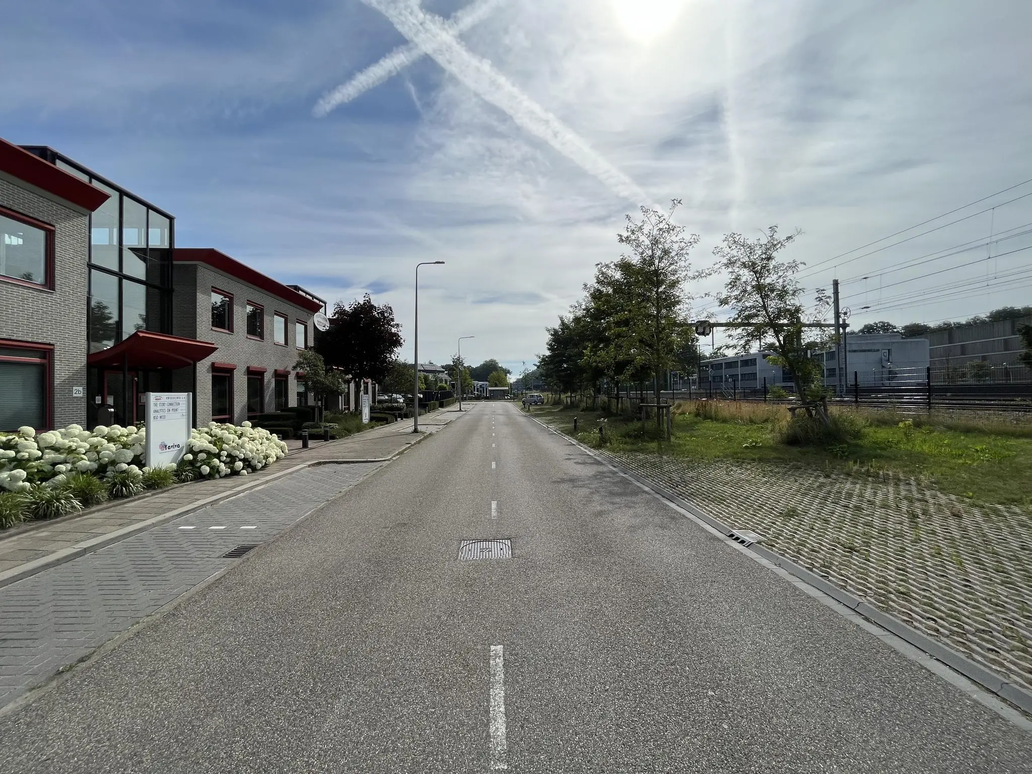 Rechthoekige straataanzicht van de Kwekerijweg met kantoorgebouwen links en een spoorlijn rechts onder een deels bewolkte lucht.