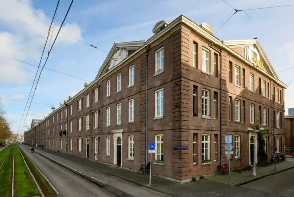 Bakstenen hoekgebouw aan de Sarphatistraat in Amsterdam met klassieke geveldetails en tramlijn op de voorgrond.