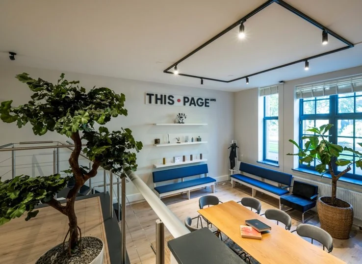 Moderne kantoorruimte aan de Sarphatistraat met blauwe zitbanken, grote kamerplanten en het logo "THIS PAGE AMS" aan de muur.