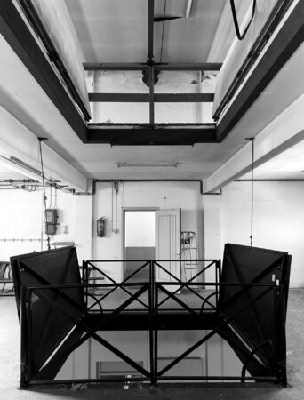 Zwart-witfoto van een industriële binnenruimte aan de Ruysdaelkade met metalen balustrades en een open vloerluik naar een lager gelegen verdieping.