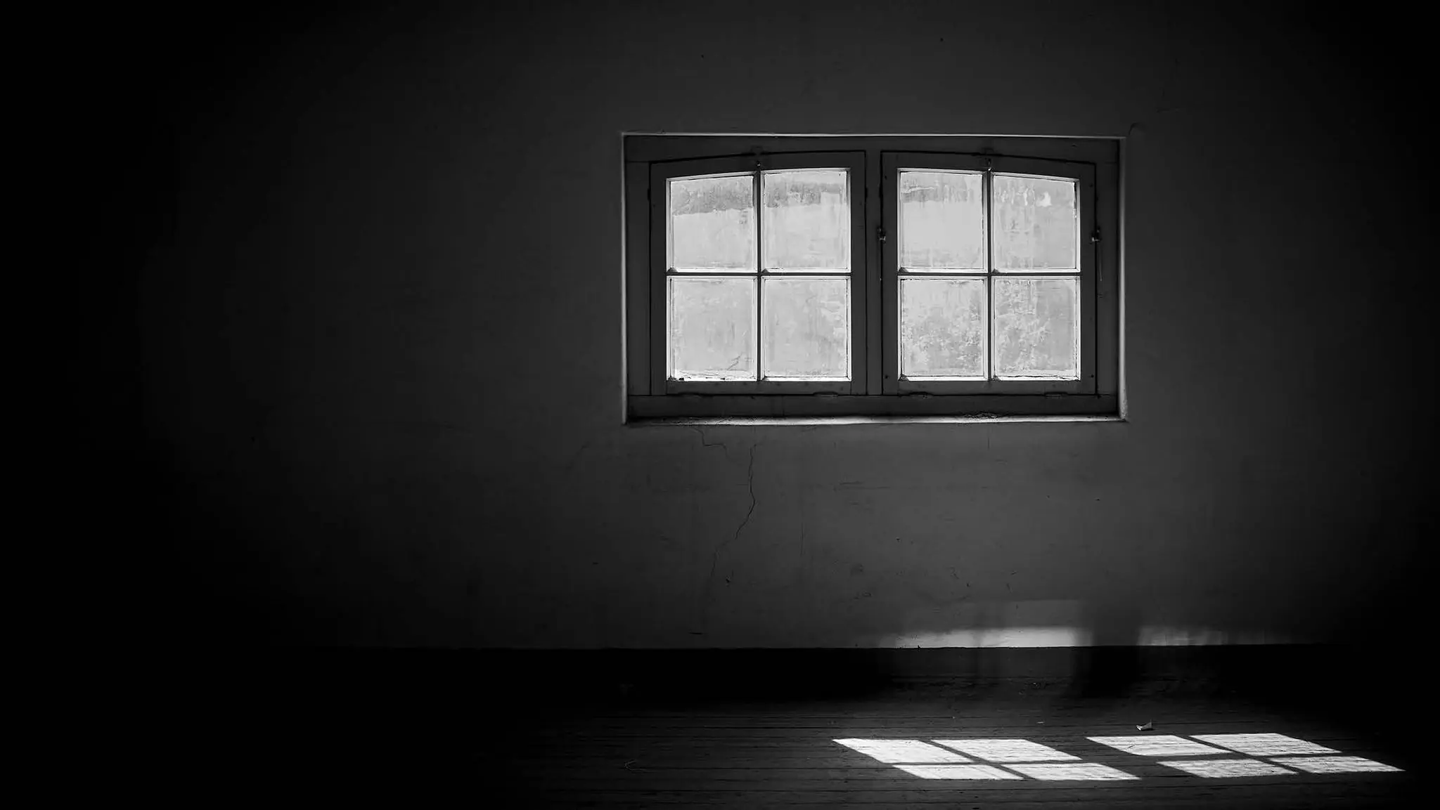 Zwart-witfoto van een leeg, donker interieur met een klassiek raam waar zonlicht door naar binnen valt.