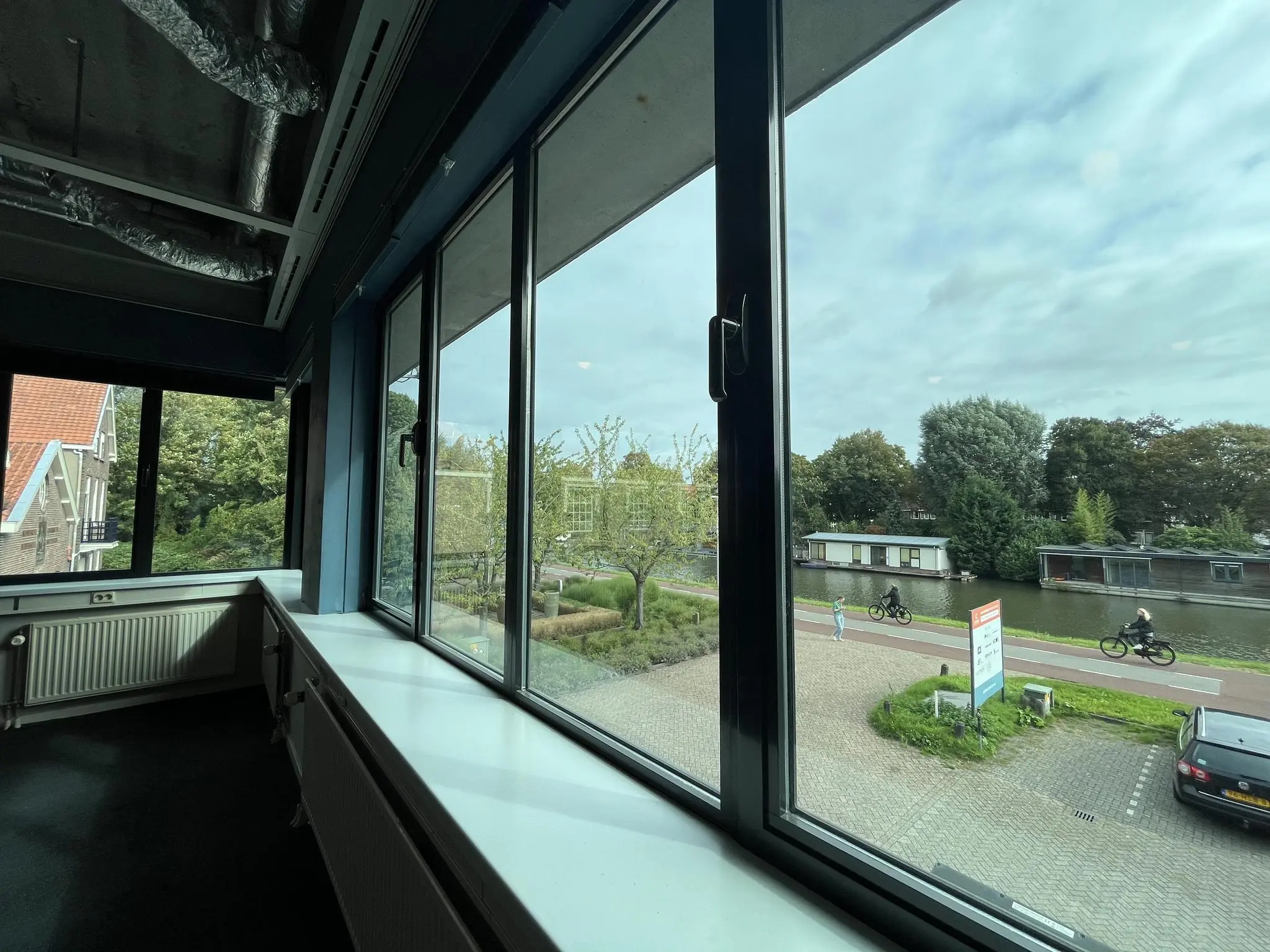 Uitzicht vanuit een kantoorraam op de Kanaalweg met fietspad, geparkeerde auto's en woonboten aan het water.