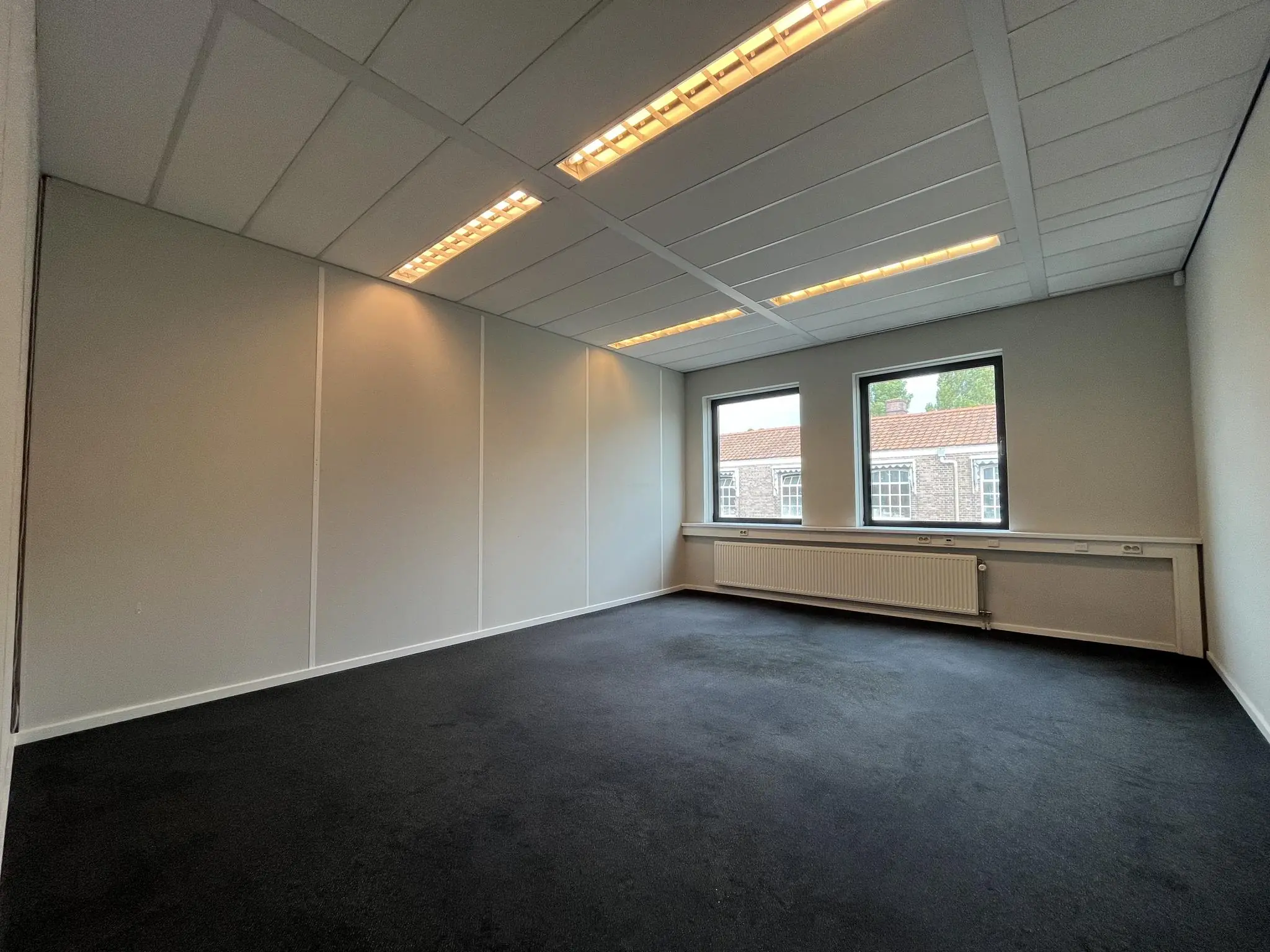 Leeg kantoorruimte aan de Kanaalweg met drie ramen, systeemplafond en donker tapijt.