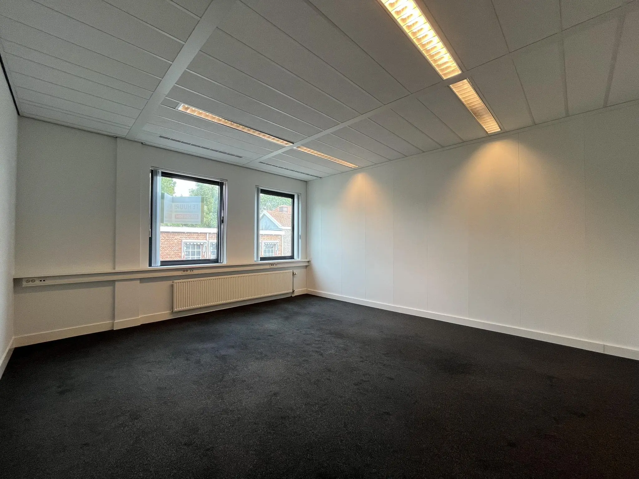 Leeg, licht kantoor met zwarte vloerbedekking en drie ramen aan de Kanaalweg.