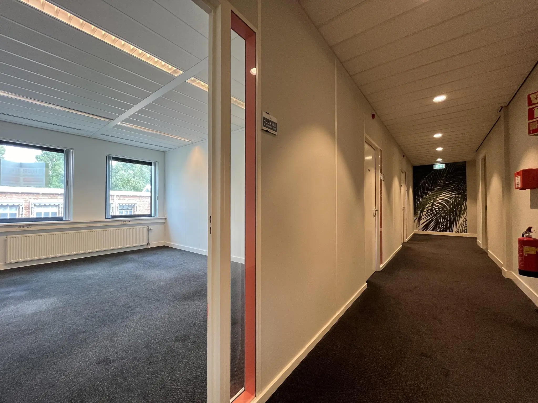 Gang in een kantoorpand aan de Kanaalweg met zicht op een leeg kantoorruimte met tapijtvloer en grote ramen.