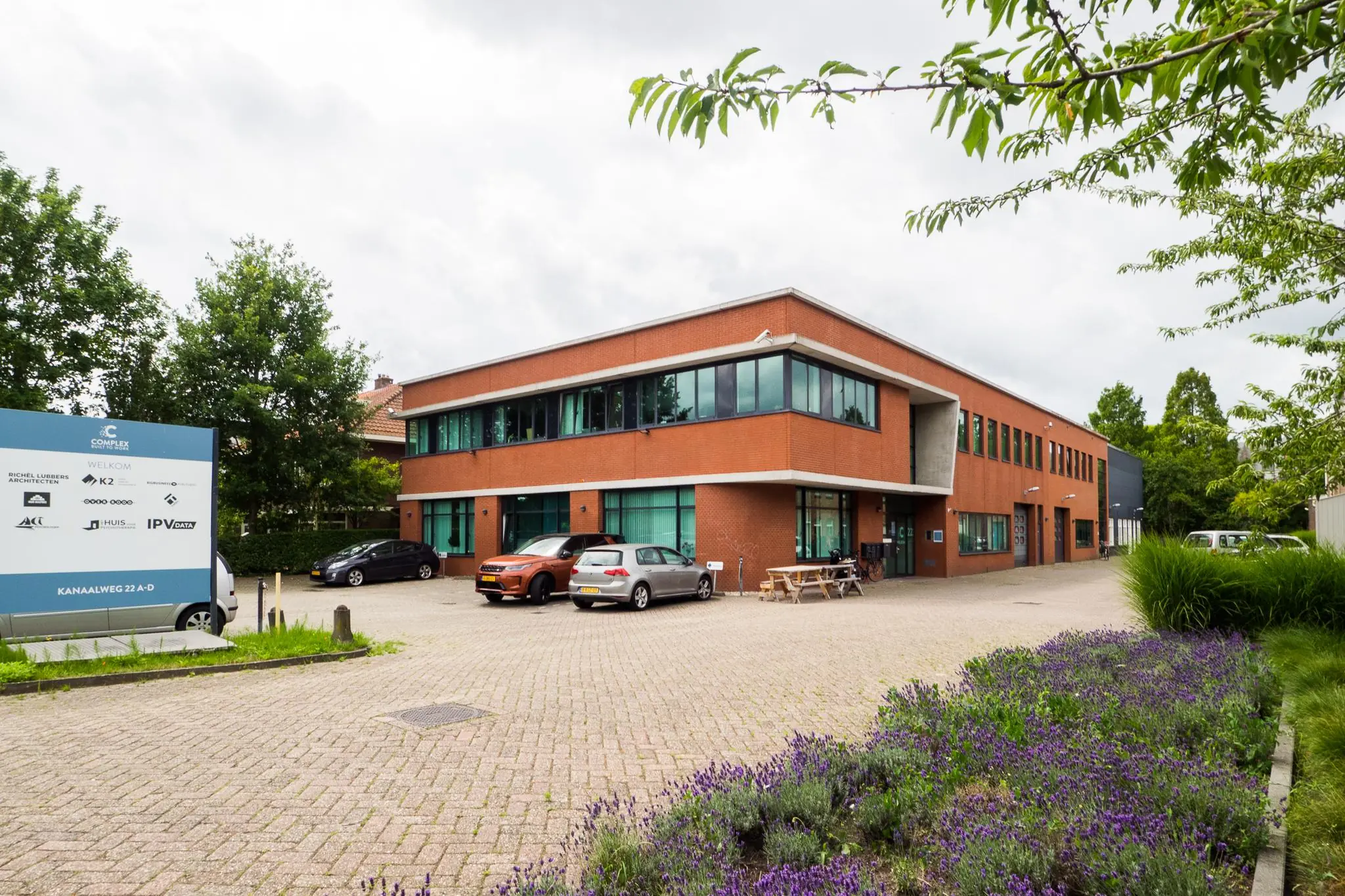 Bedrijfsverzamelgebouw aan de Kanaalweg 22 met parkeerplaatsen, tuin met lavendel en een informatiebord met bedrijfsnamen.