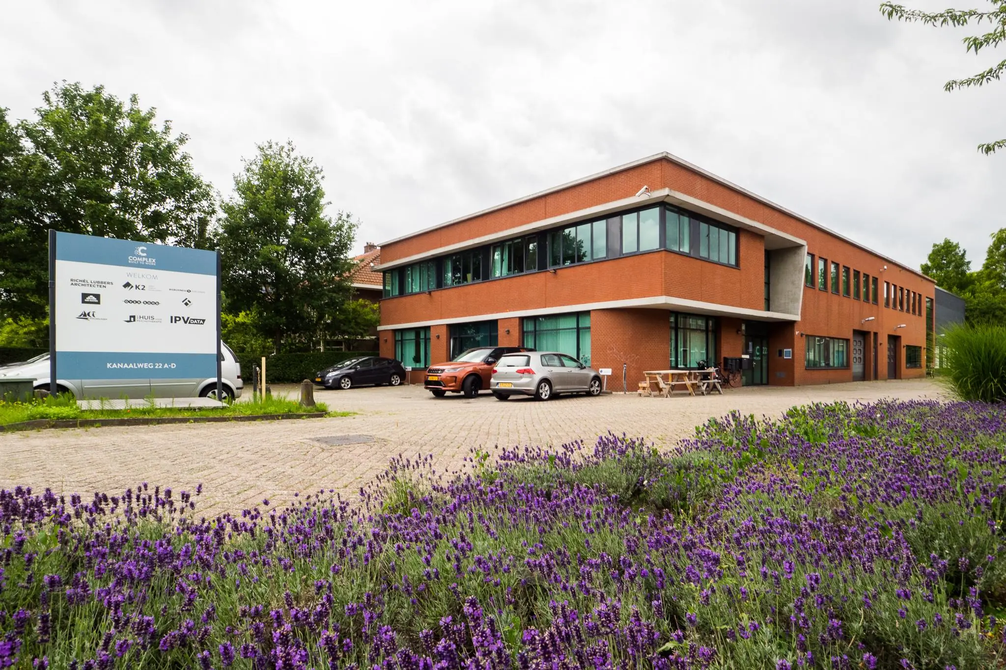 Modern kantoorpand aan de Kanaalweg 22 met parkeerplaatsen, een informatiebord en bloeiende paarse lavendel op de voorgrond.