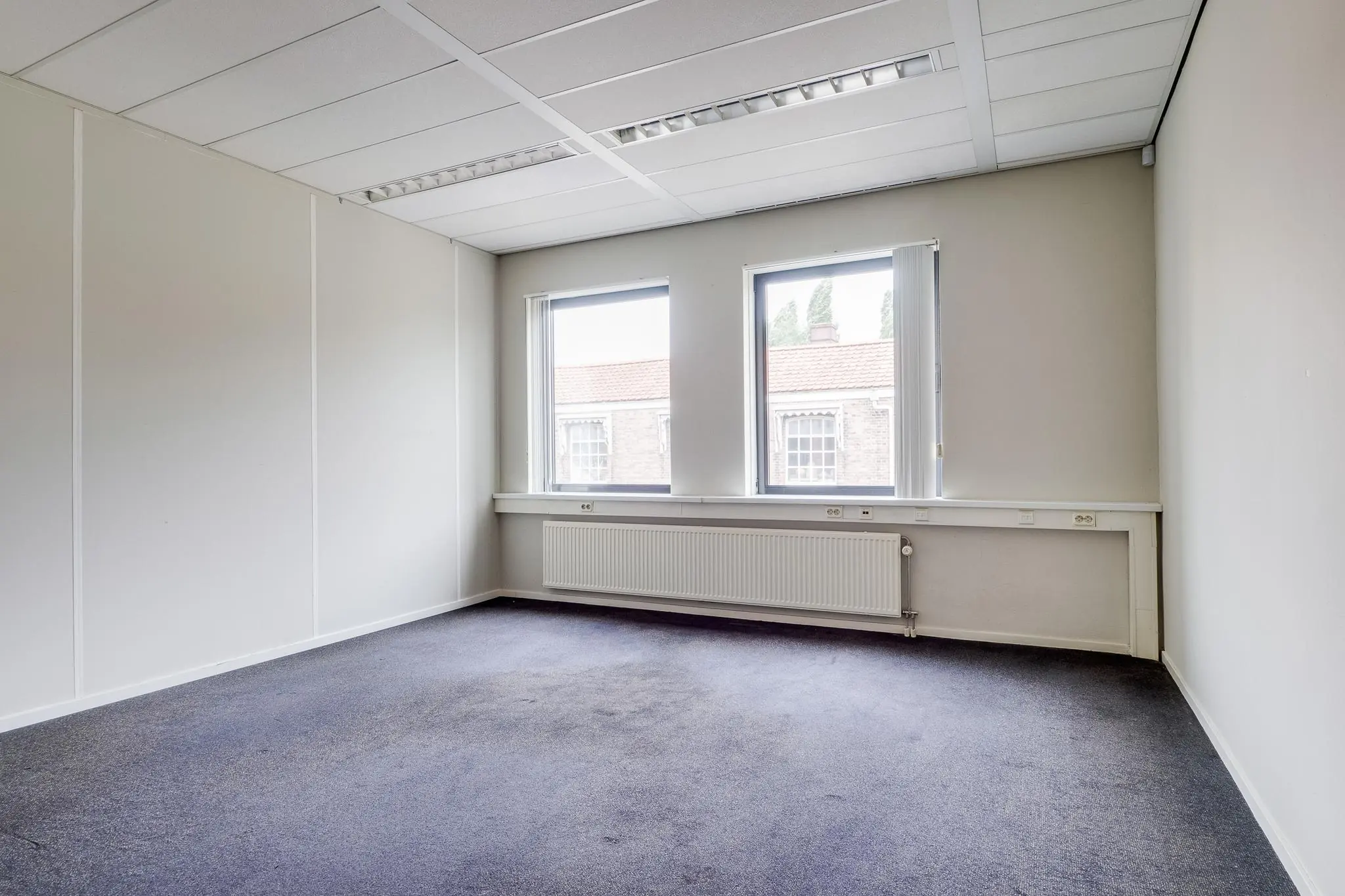 Leeg kantoor met lichtgrijze muren, donkerblauwe vloerbedekking en twee ramen aan de Kanaalweg.