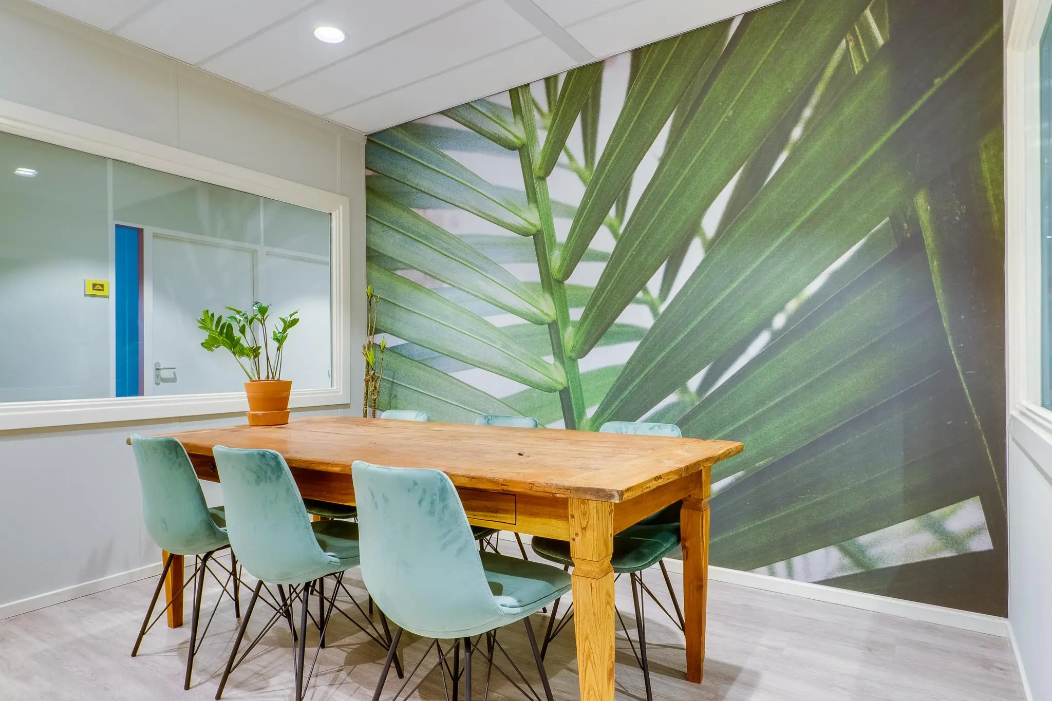 Vergaderruimte met houten tafel, zes mintgroene stoelen en een wand met groot palmblaadmotief.