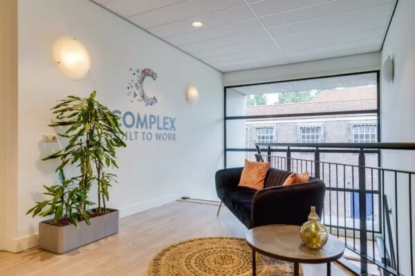 Moderne wachtruimte met een zwarte bank, kamerplant, ronde tafel en het logo van "Complex Built to Work" op de muur.