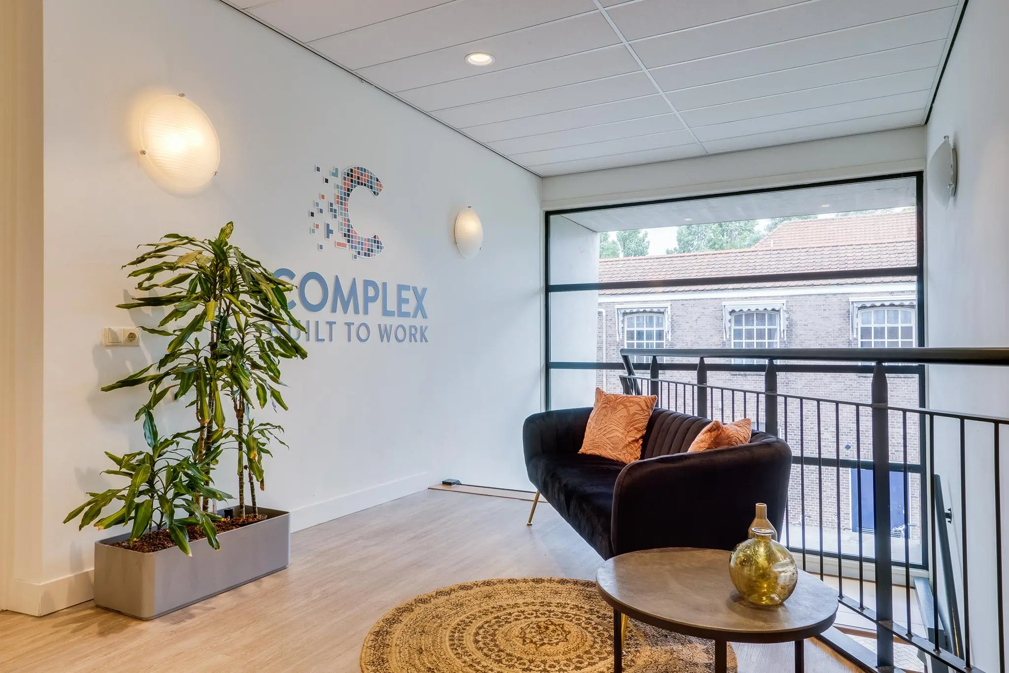 Moderne wachtruimte met een zwarte bank, kamerplant, ronde tafel en het logo van "Complex Built to Work" op de muur.