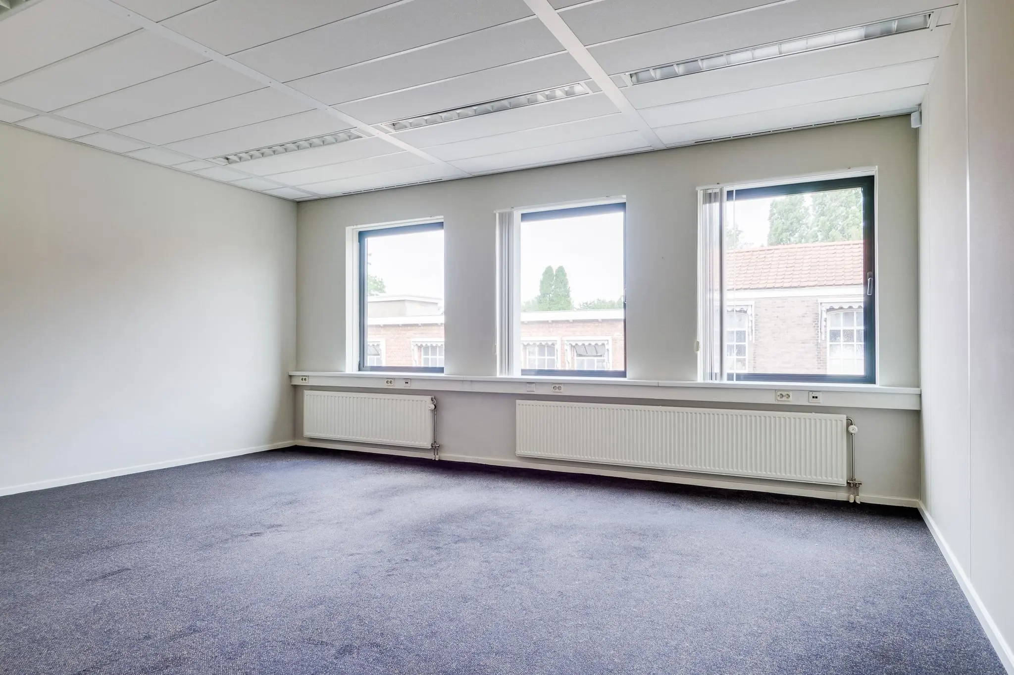 Leeg kantoor met drie ramen aan de Kanaalweg, voorzien van wit plafond, radiatoren en blauw tapijt.