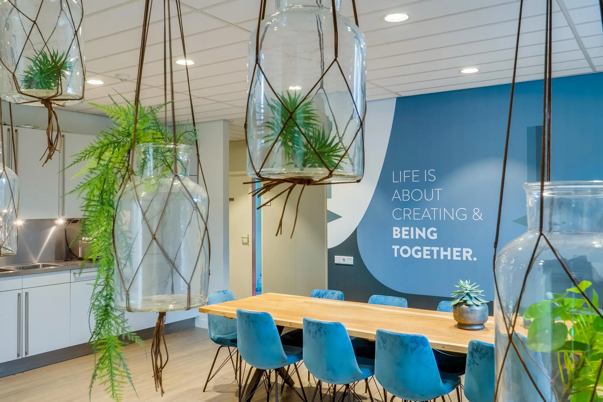 Moderne vergaderruimte aan de Kanaalweg met houten tafel, blauwe stoelen, hangende glazen plantenpotten en inspirerende muurtekst.