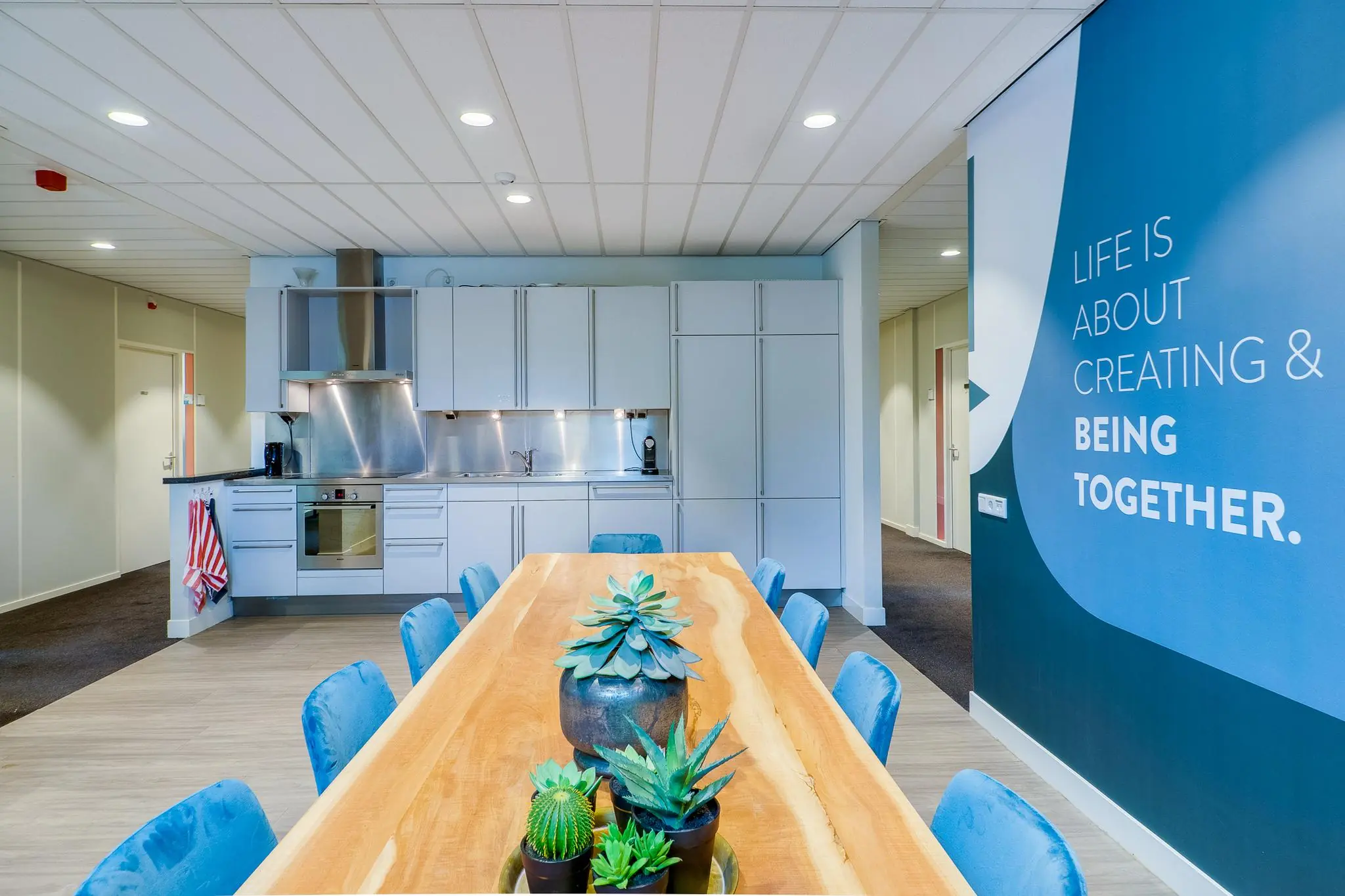 Moderne gezamenlijke keuken met een lange houten eettafel, blauwe stoelen en een motiverende tekst op de wand.
