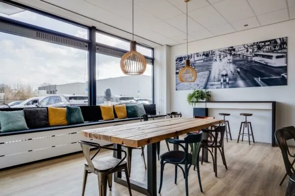 Moderne bedrijfsruimte met een houten vergadertafel, industriële stoelen, kleurrijke kussens bij het raam en een zwart-wit stadsfoto aan de muur.