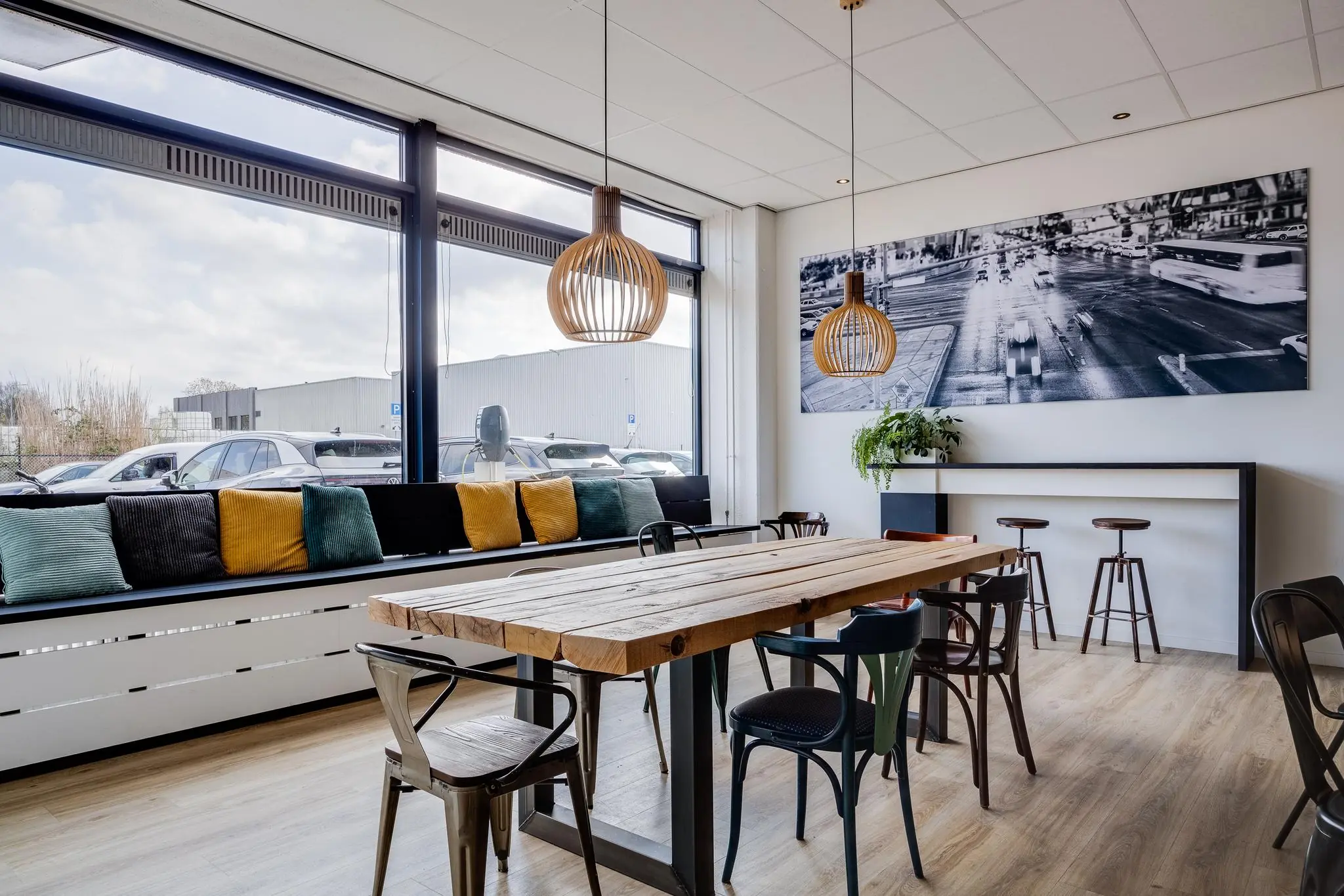 Moderne bedrijfsruimte met een houten vergadertafel, industriële stoelen, kleurrijke kussens bij het raam en een zwart-wit stadsfoto aan de muur.