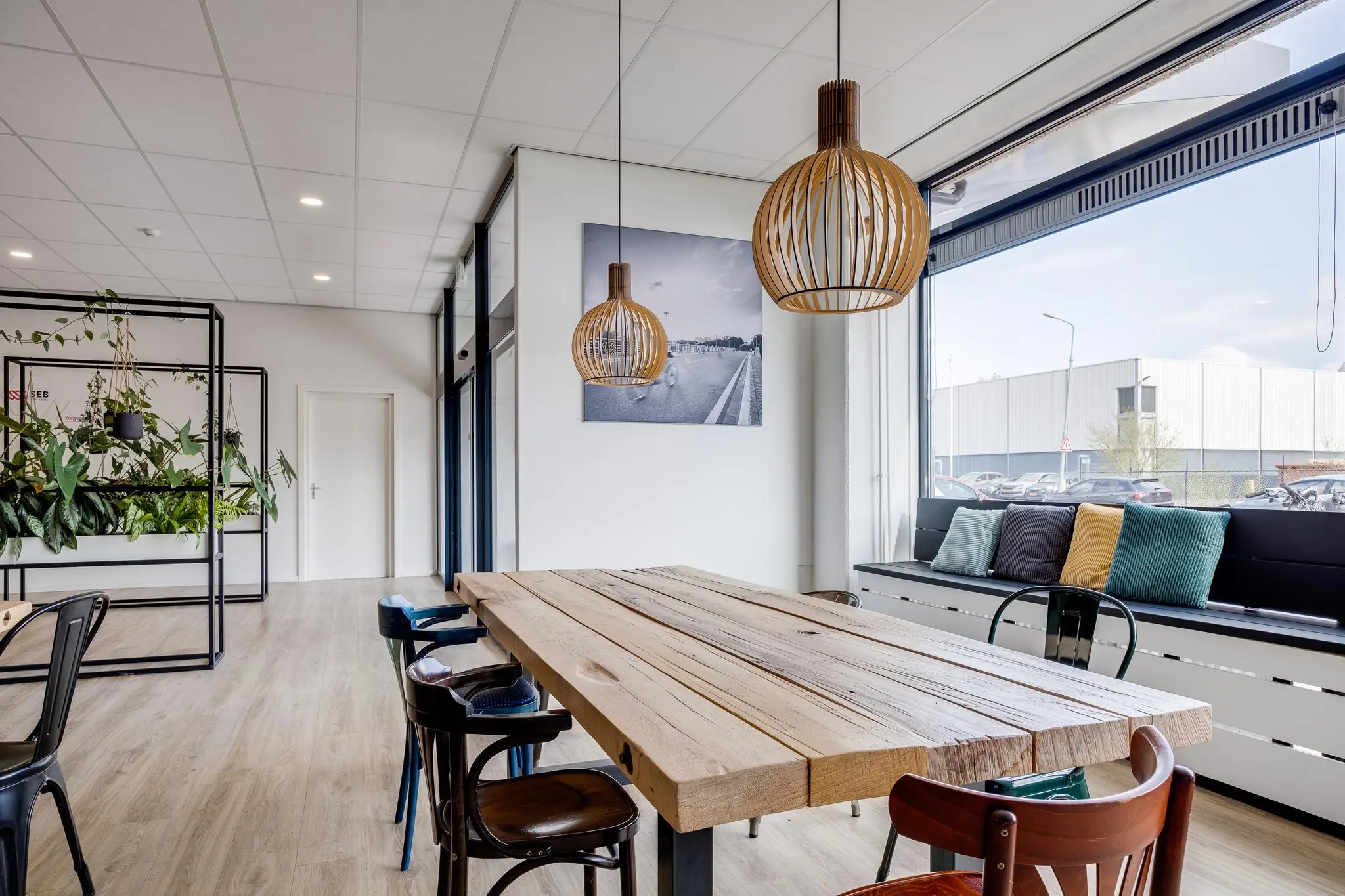 Moderne, lichte kantineruimte aan de Zonnebaan met houten tafel, hanglampen, plantenwand en groot raam met uitzicht op parkeerterrein.