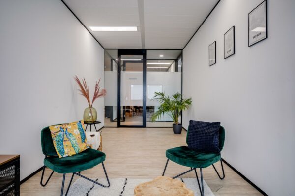 Moderne wachtruimte met twee groene stoelen, plantendecoratie en zicht op vergaderruimte via glazen deur.