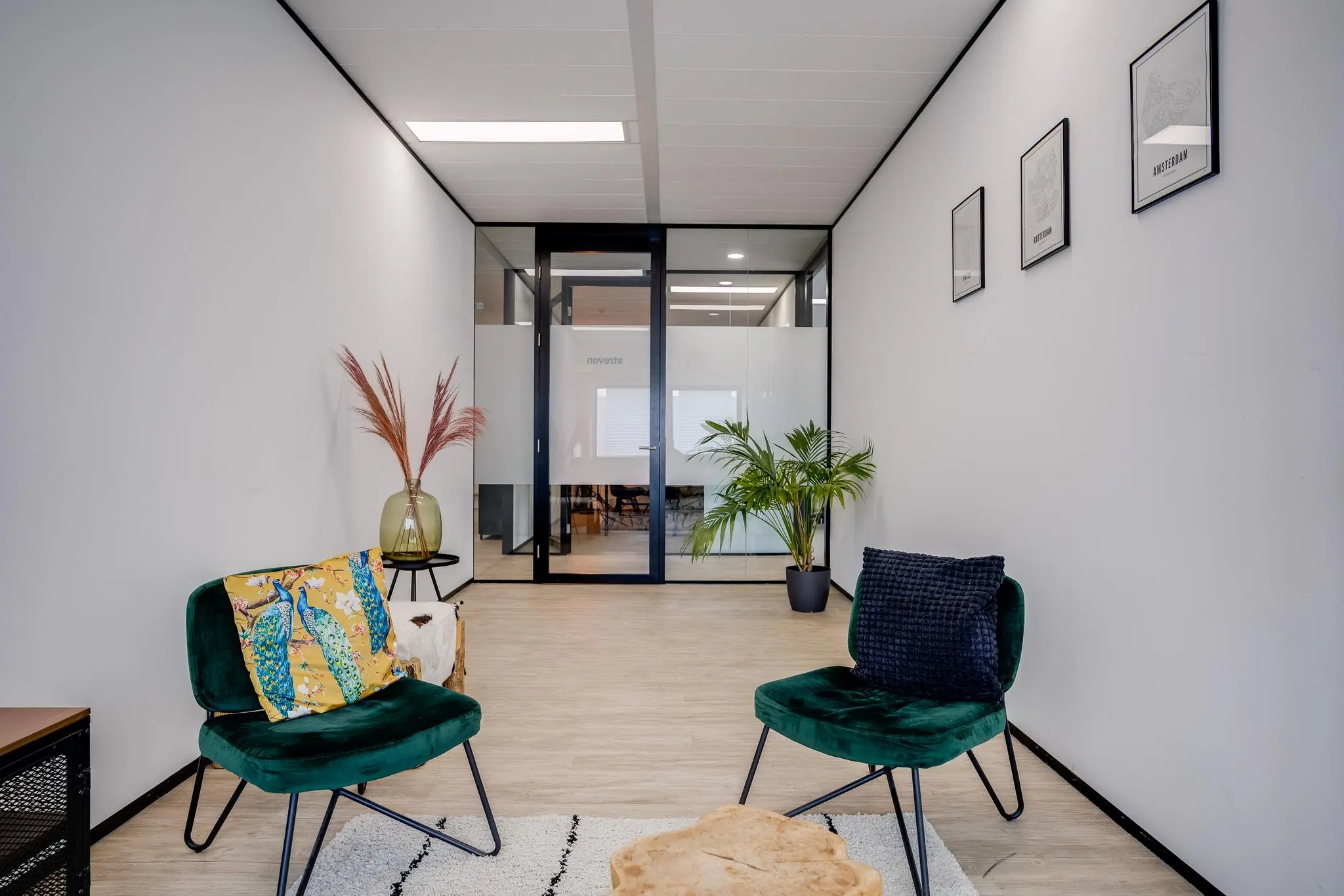 Moderne wachtruimte met twee groene stoelen, plantendecoratie en zicht op vergaderruimte via glazen deur.