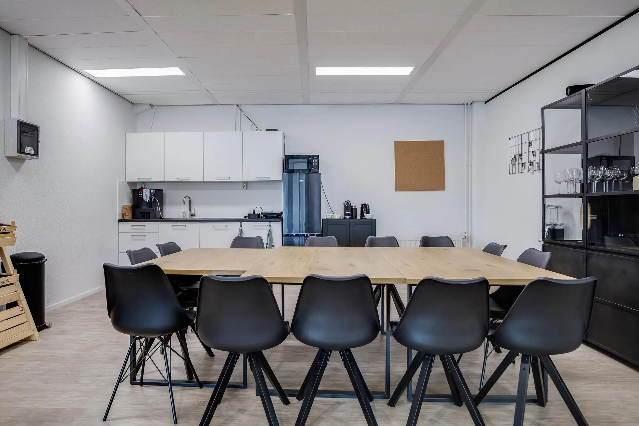 Vergaderruimte met lange houten tafel, zwarte stoelen en een kitchenette op de achtergrond.