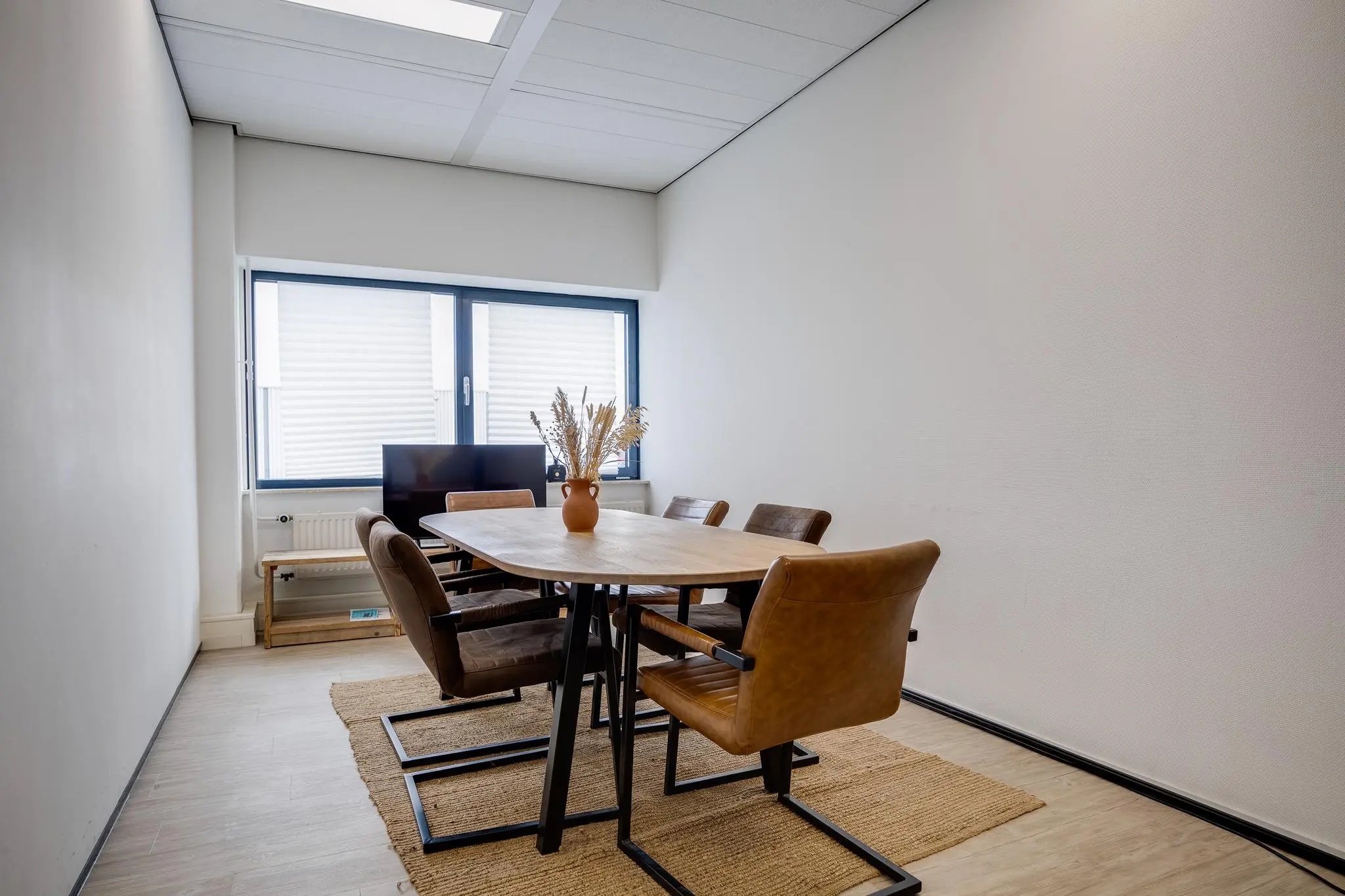 Vergaderruimte met houten tafel, zes leren stoelen en een vaas met droogbloemen bij het raam.