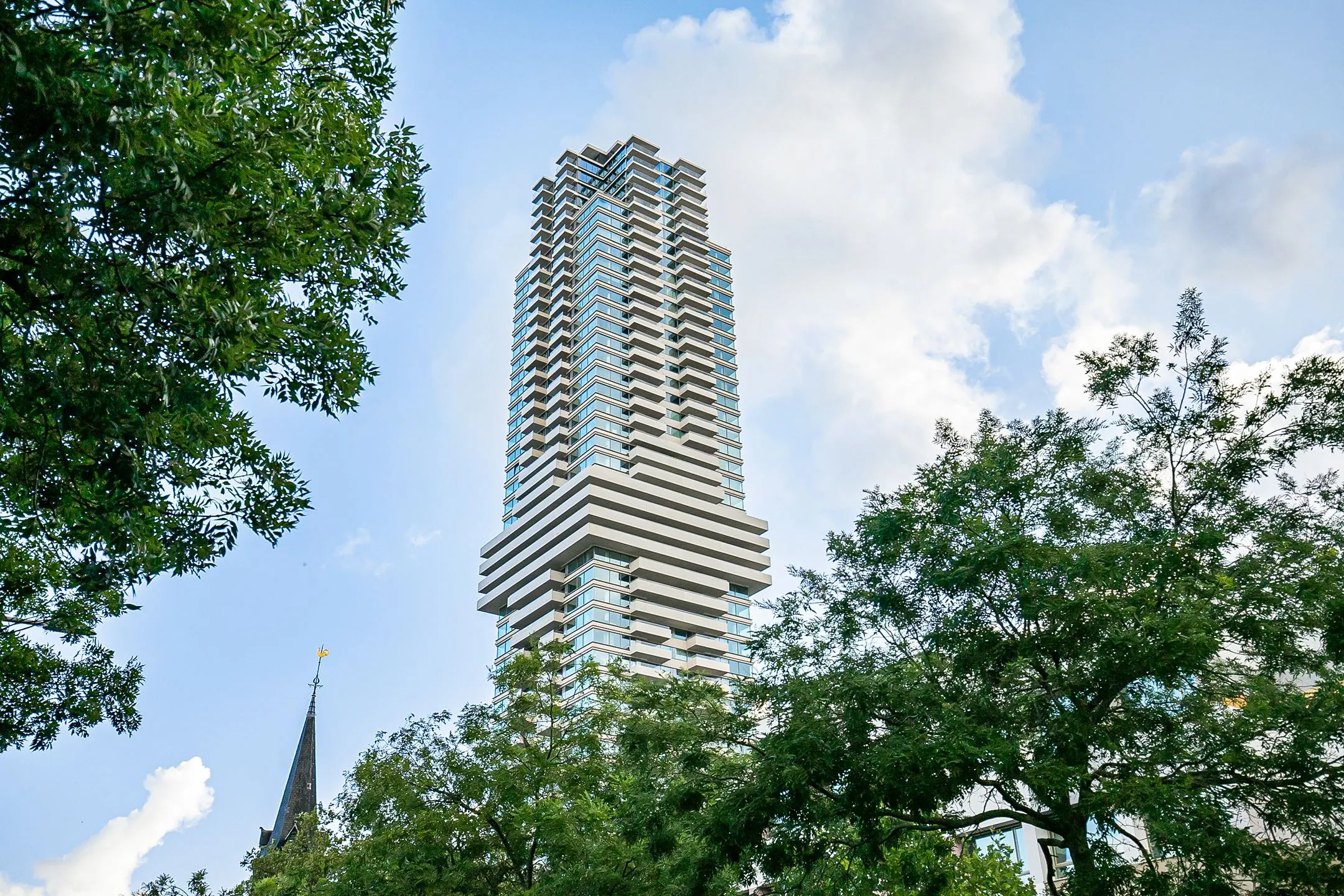 Modern high-rise building "De Baan" in Rotterdam viewed from below, framed by green trees and a partly cloudy sky.
