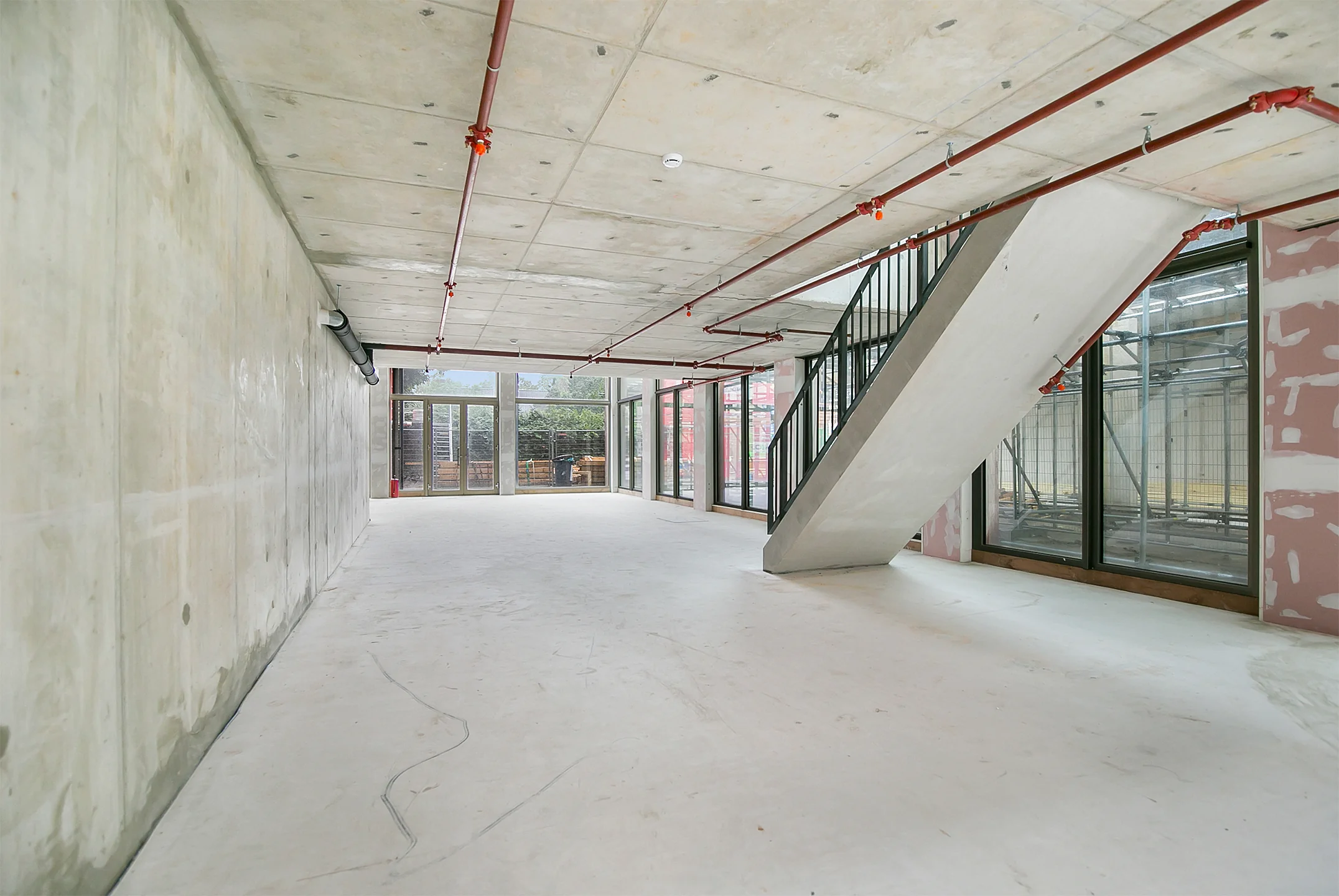 Empty concrete interior of a commercial or residential space under construction with visible piping and a staircase.