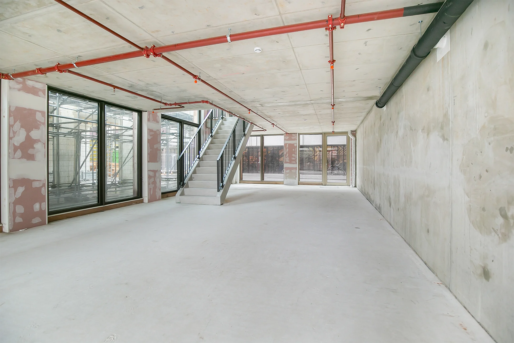 Empty commercial space under construction with concrete floors, large windows, and a central staircase.