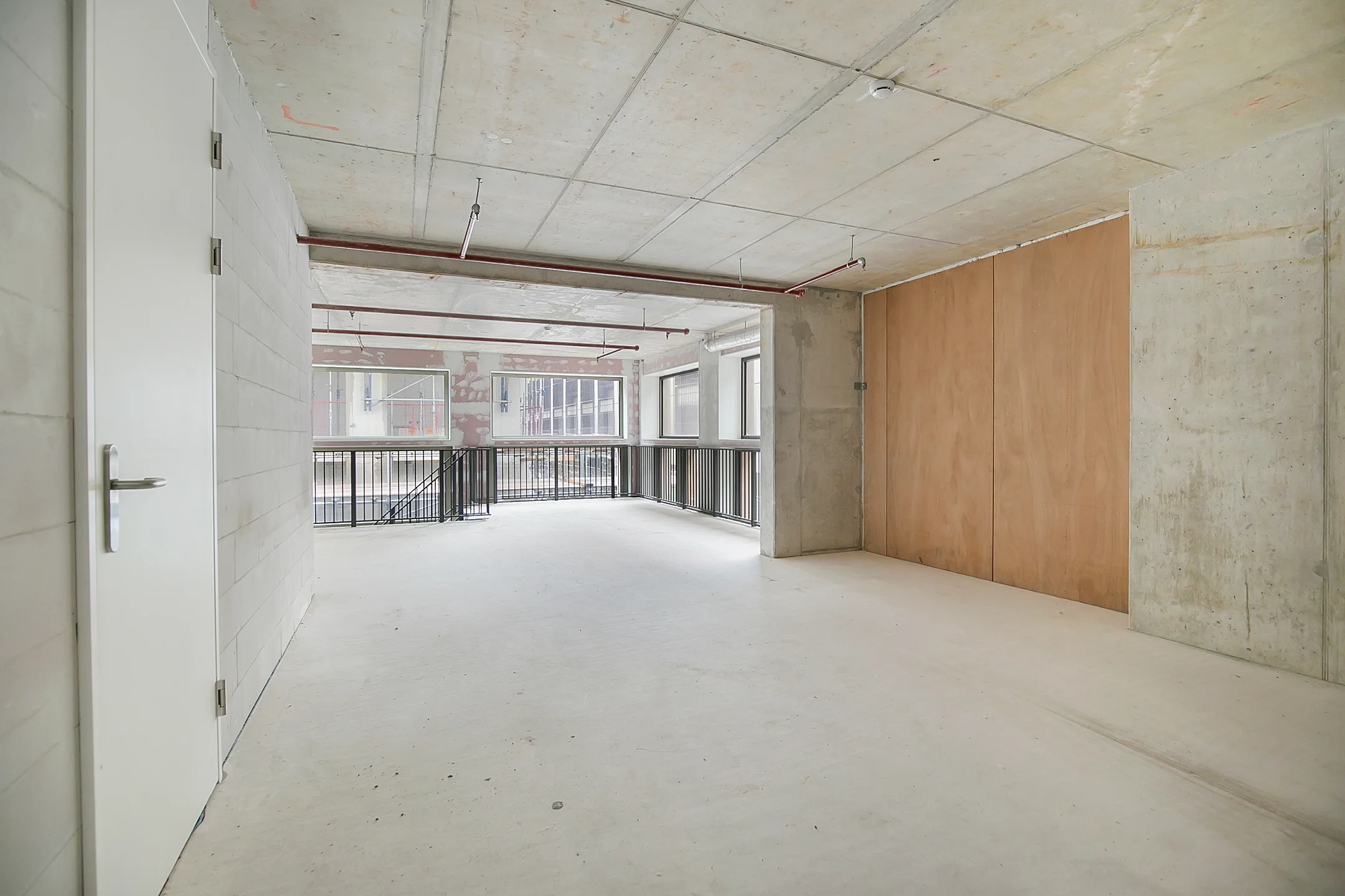 Empty industrial-style interior with concrete walls, exposed ceiling pipes, and large windows overlooking a lower level.