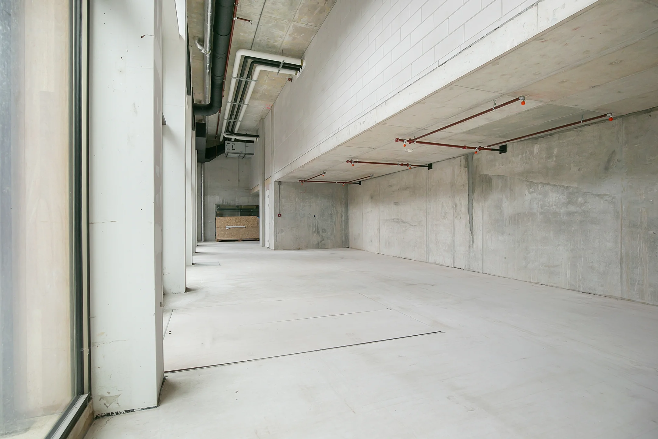 Empty industrial-style commercial space with concrete walls, exposed pipes, and large windows.