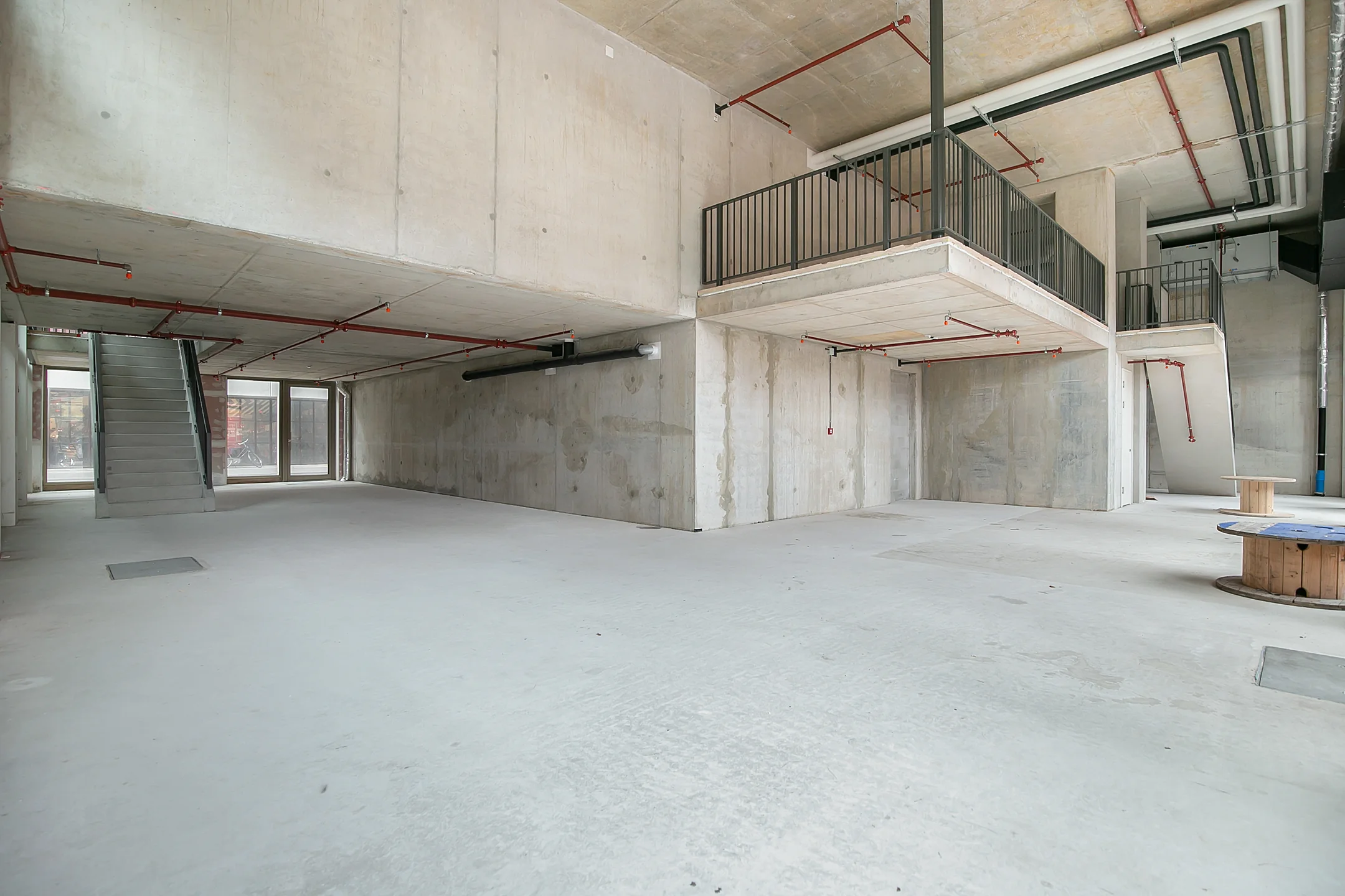 Spacious unfinished concrete commercial interior with exposed pipes, stairs, and large windows.