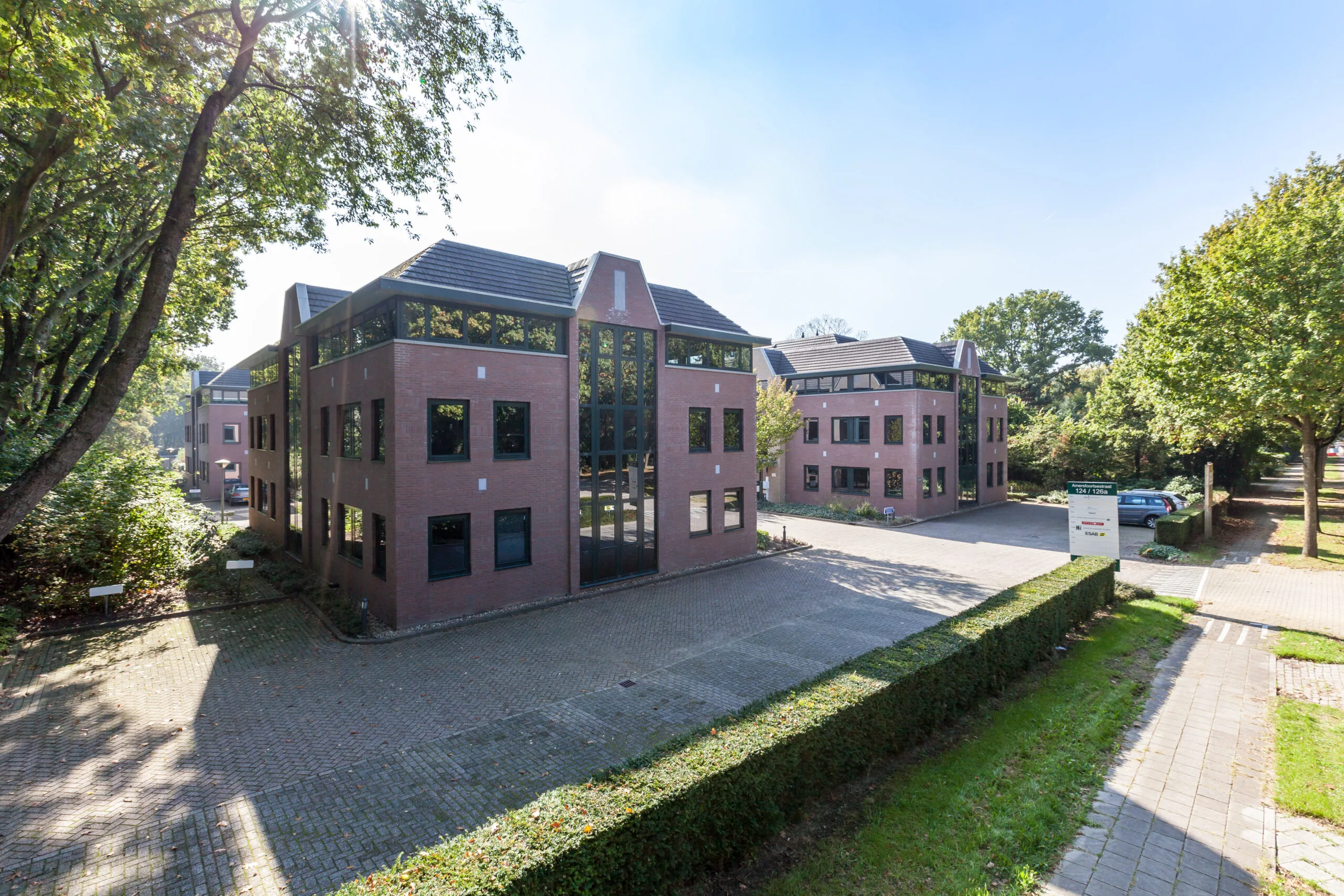 Twee moderne kantoorgebouwen van rood baksteen met donkere raamkozijnen, omgeven door groen en een bestrate parkeerplaats.