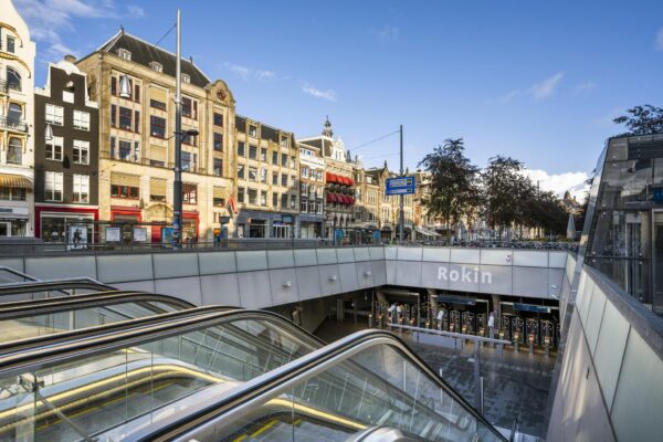 Ingang van metrostation Rokin in Amsterdam met roltrappen en historische gebouwen op de achtergrond.