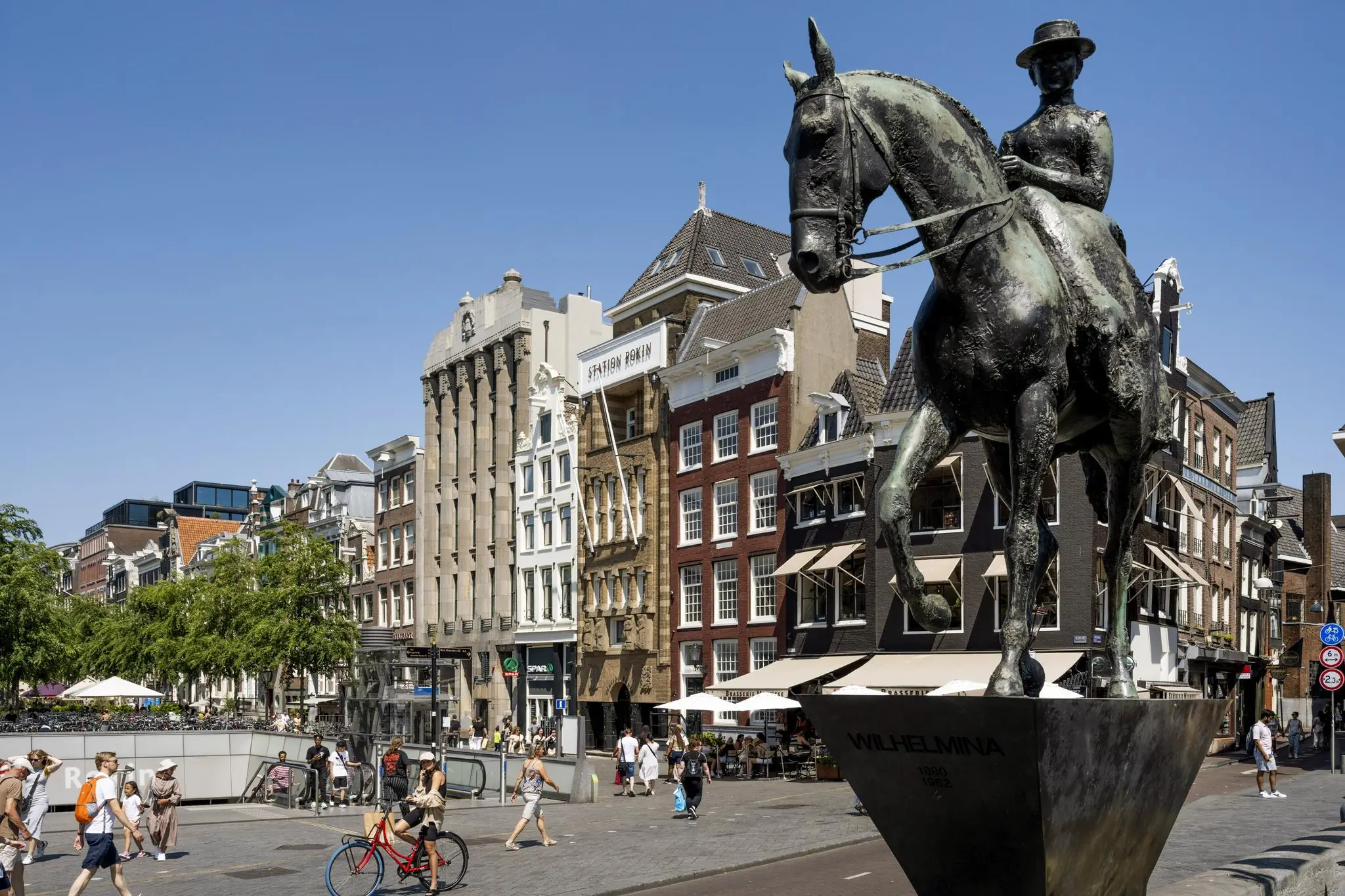 Standbeeld van koningin Wilhelmina te paard op het Rokin in Amsterdam, met historische gevels en Station Rokin op de achtergrond.