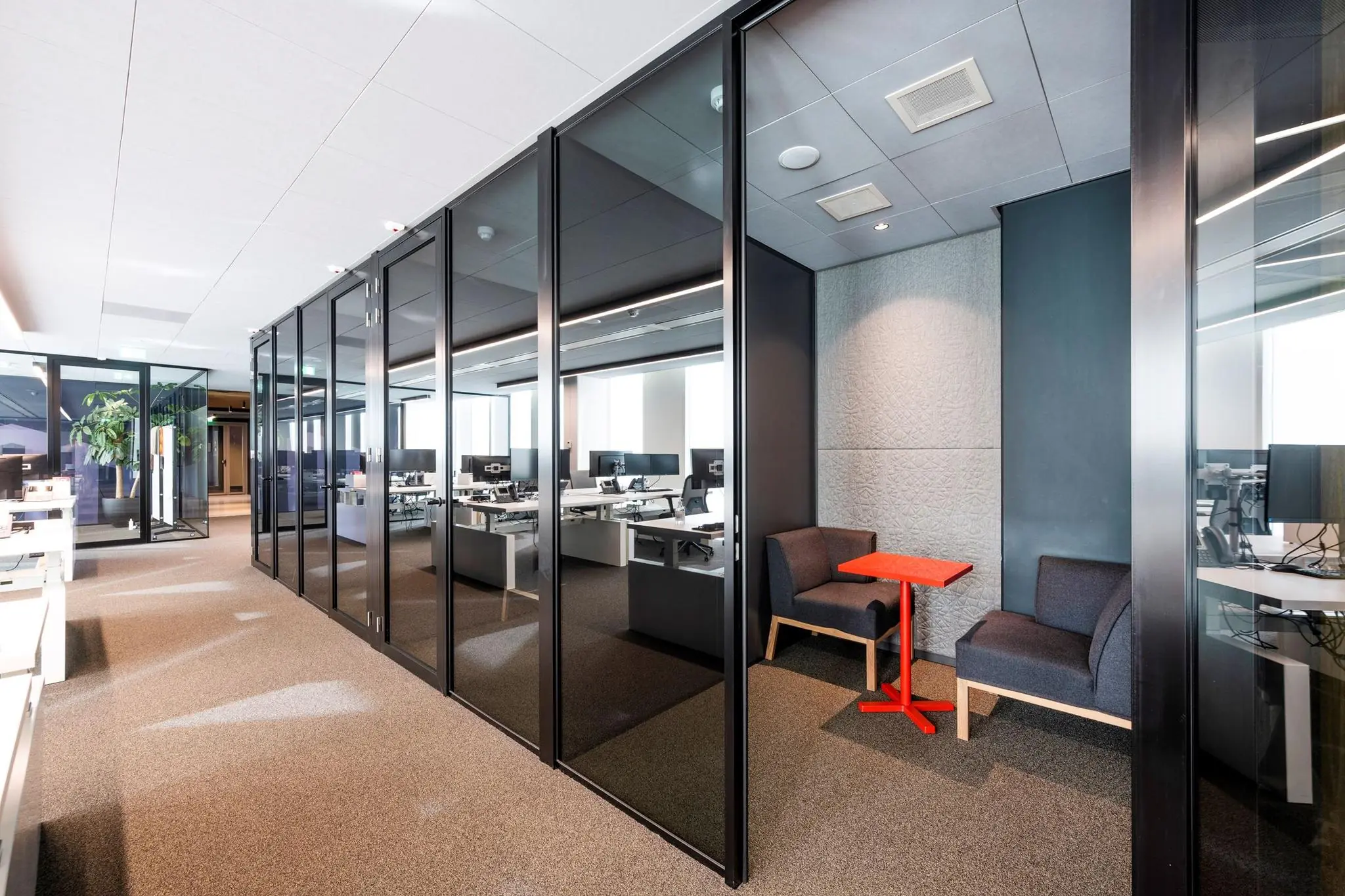 Moderne kantoorruimte in gebouw Hoekenrode met glazen vergaderruimtes, werkplekken en een zitje met rood tafeltje.