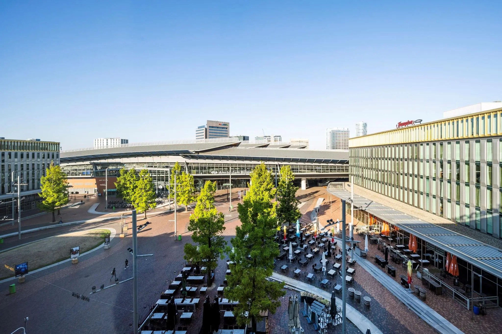 Station Amsterdam Bijlmer ArenA met omliggende horecaterrassen en moderne gebouwen op een zonnige dag.