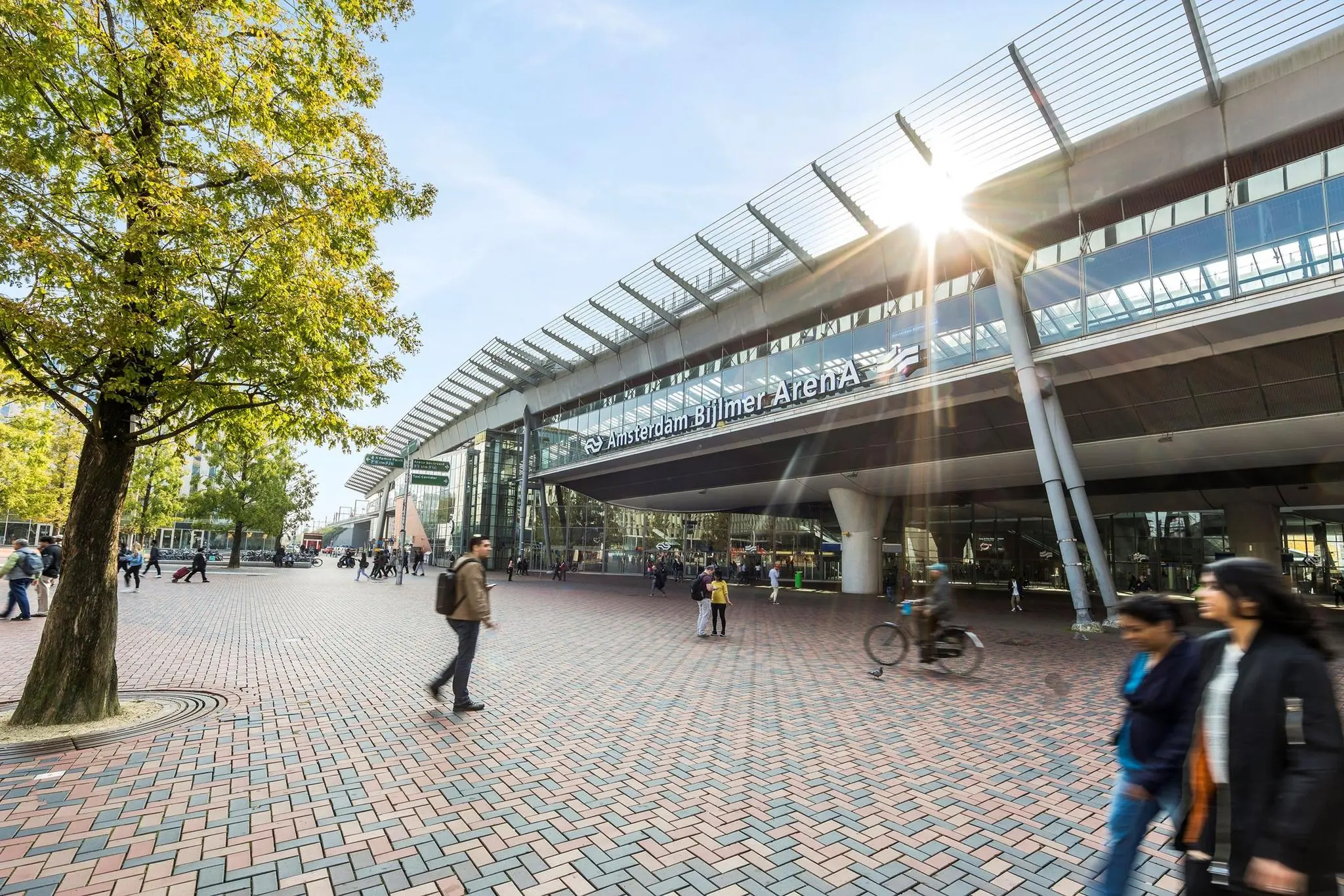 Voorzijde van station Amsterdam Bijlmer ArenA met passerende voetgangers en fietsers op het plein.