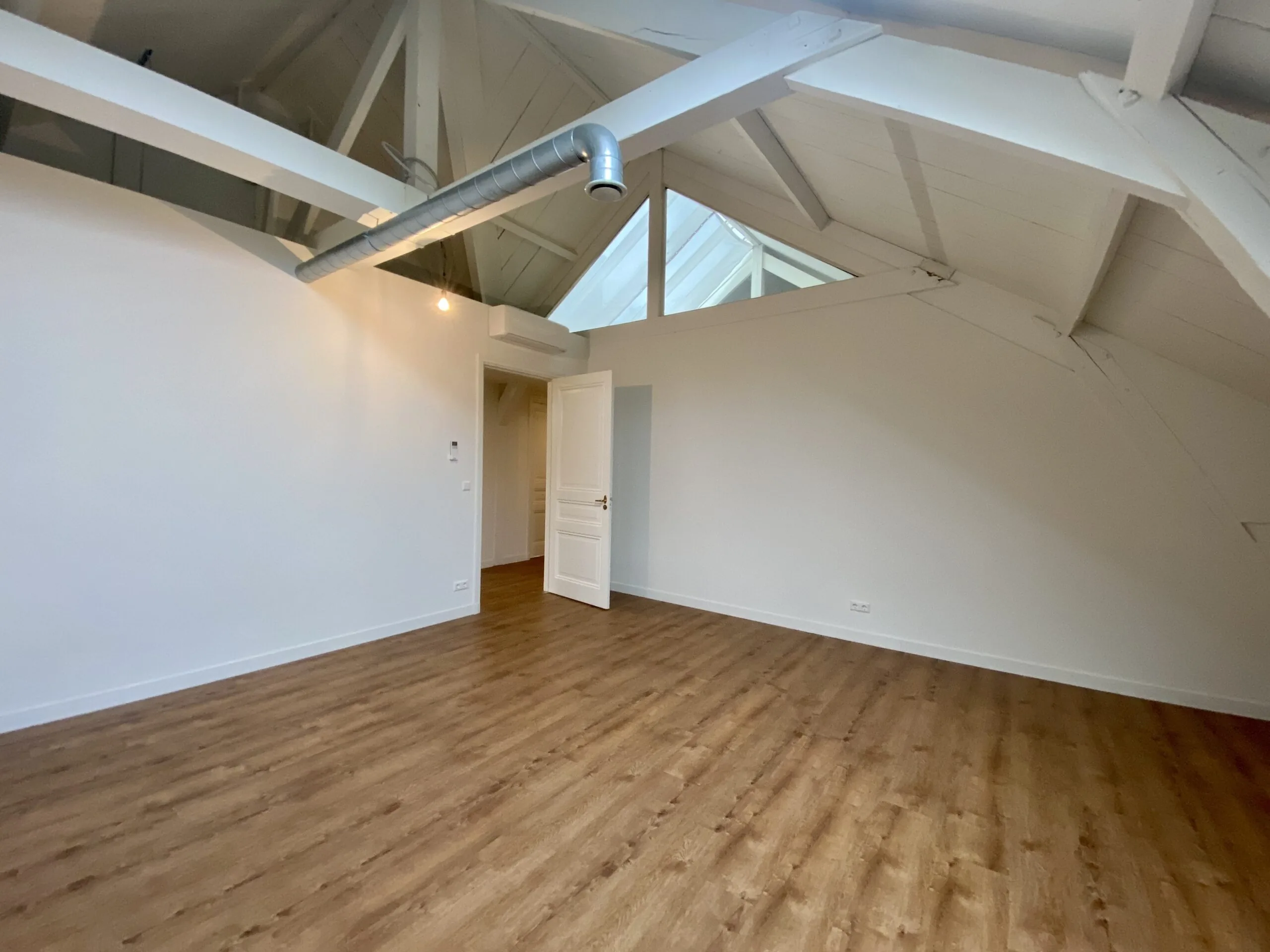 Ruime zolderkamer met houten vloer, witte balkenplafond en openstaande deur.