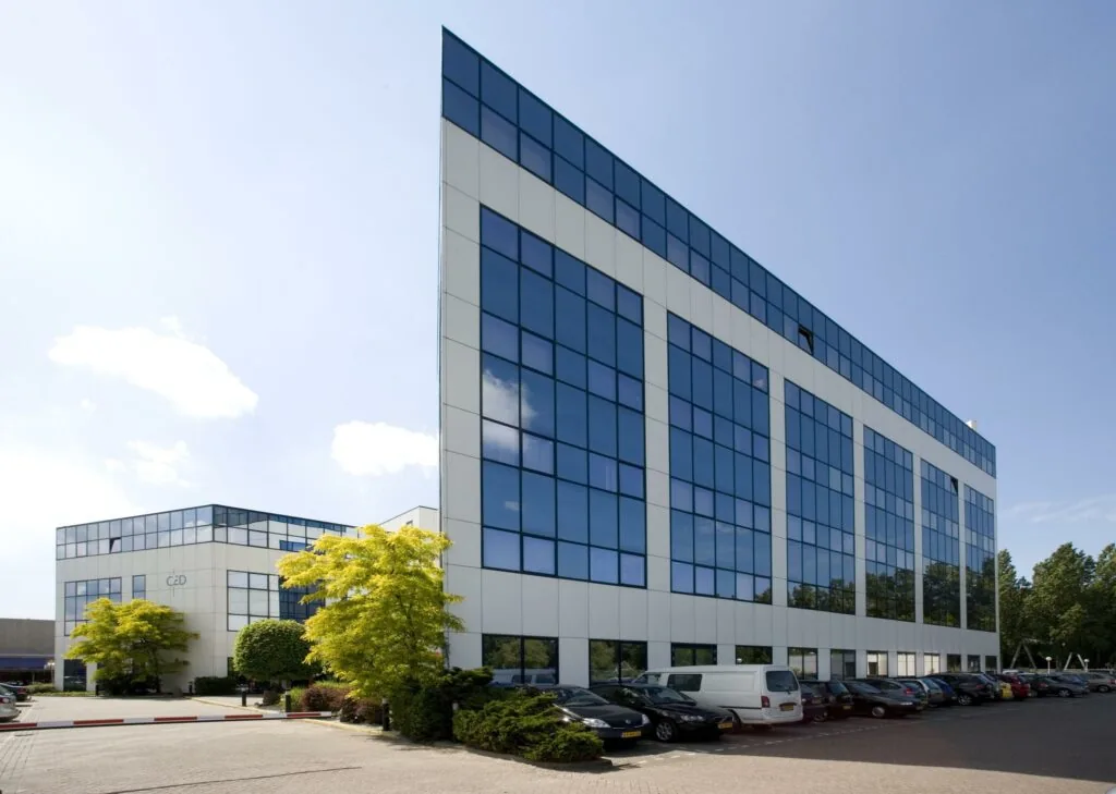 Modern kantoorgebouw aan de Rietbaan met spiegelende ramen en parkeerplaats op de voorgrond.