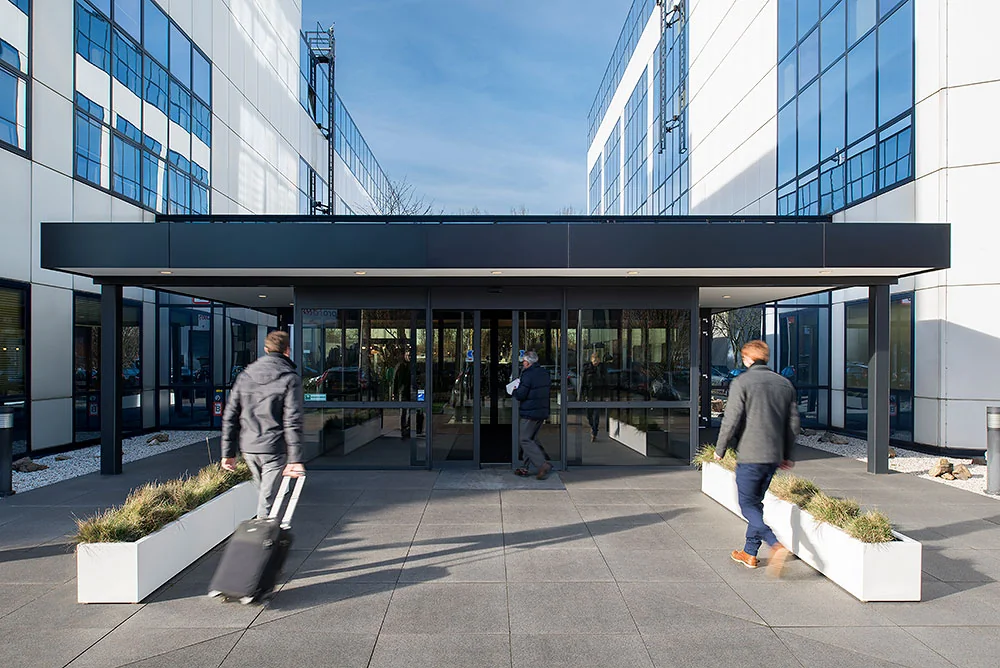 Ingang van een modern kantoorgebouw aan de Rietbaan met mensen die naar binnen lopen.