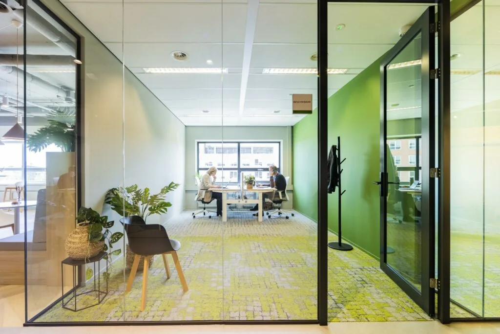 Twee mensen werken tegenover elkaar in een modern, licht kantoor aan de Rietbaan met groene muren en glazen wanden.