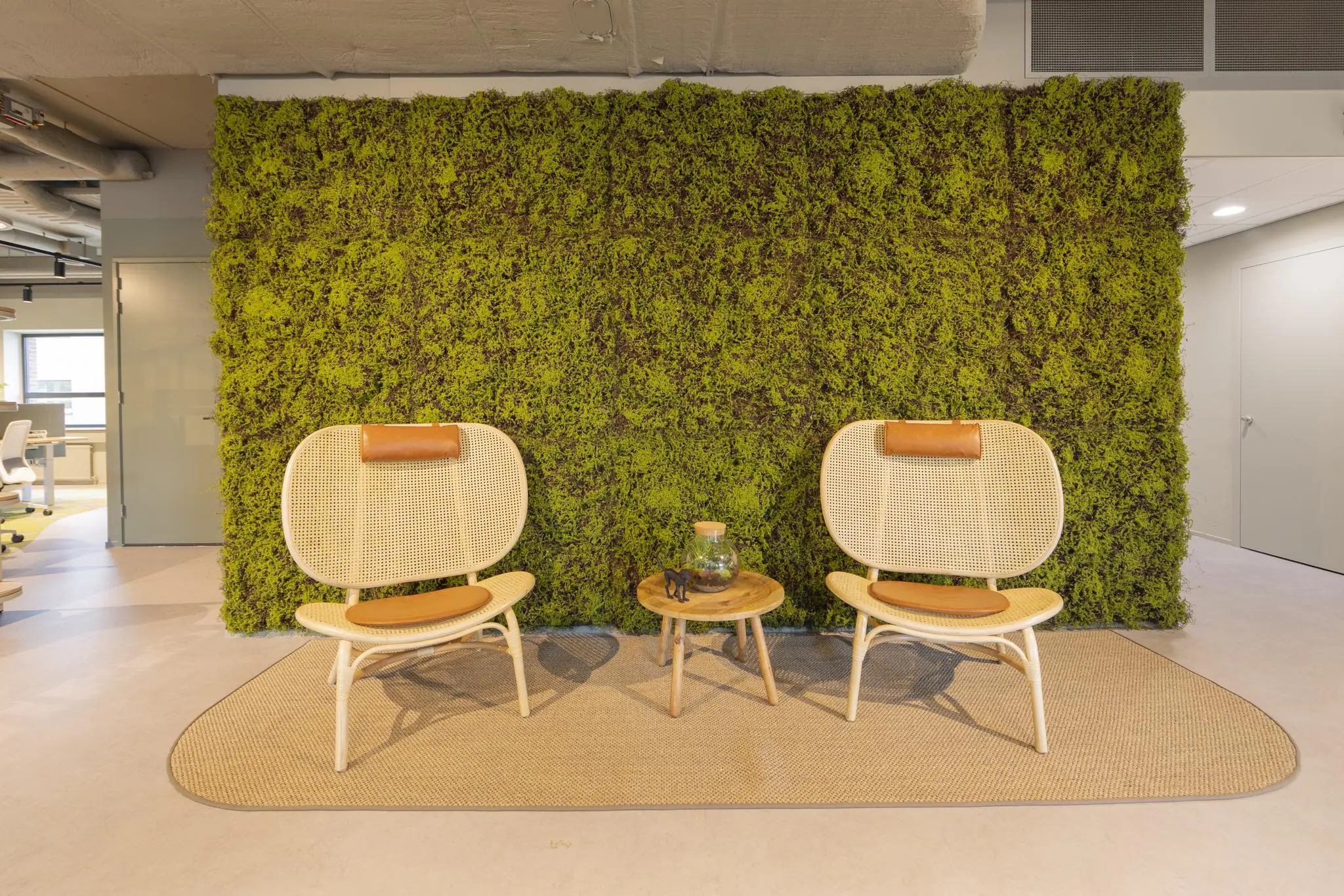 Twee rotan stoelen met lederen accenten staan op een vloerkleed voor een groene moswand in een modern kantoorinterieur.